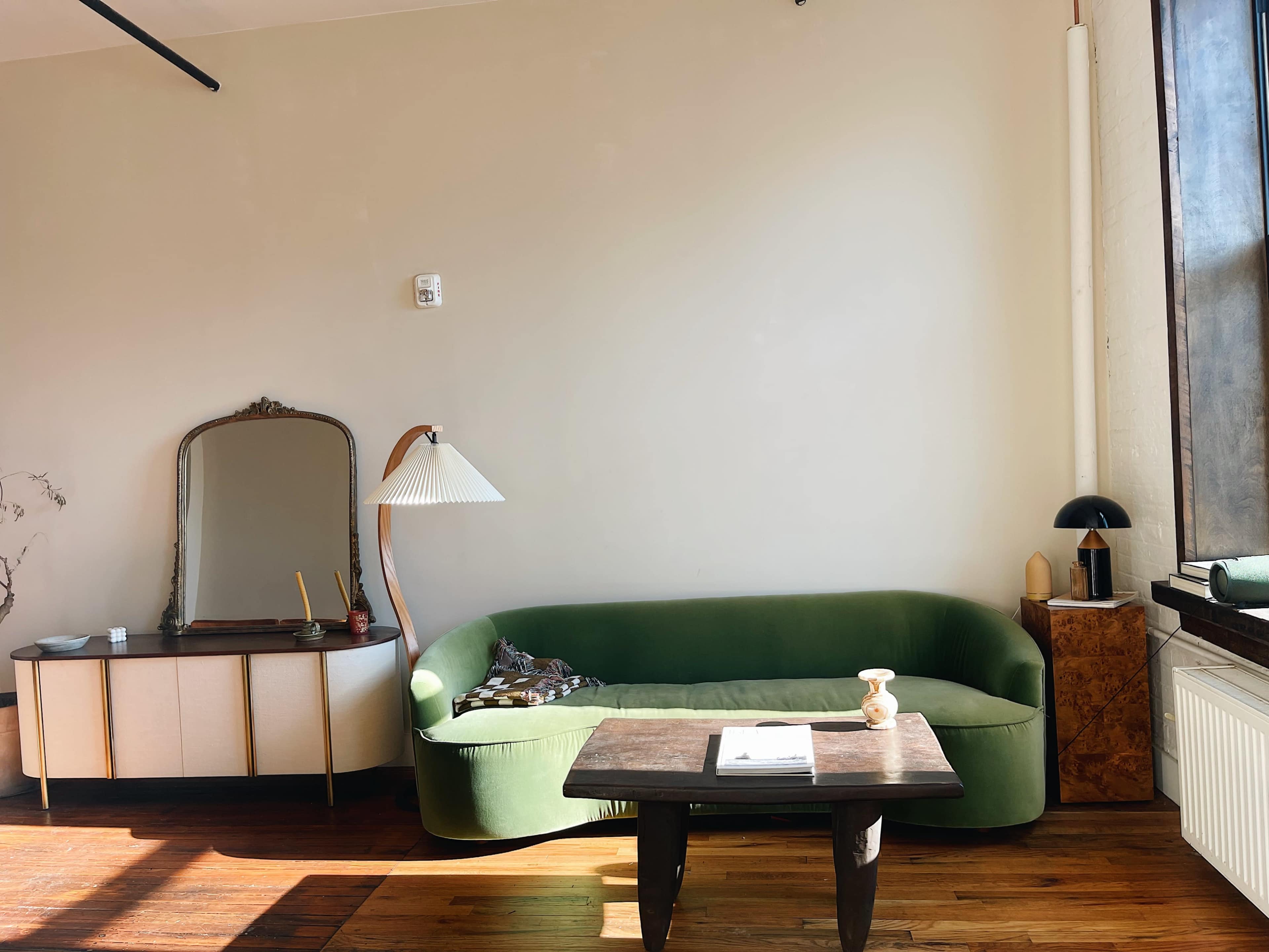 A simple living space with a green sofa, a wooden table, and a vintage mirror against a light-colored wall.