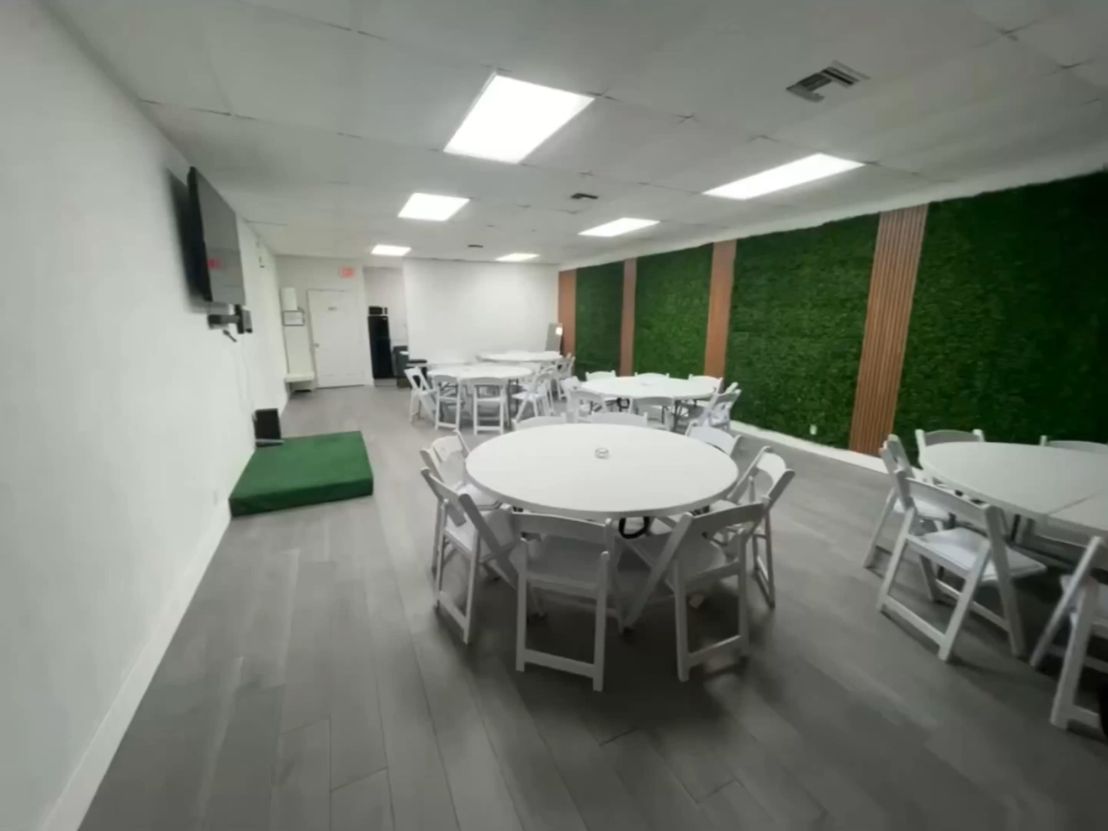 The image shows a modern, sparsely furnished event space with round tables arranged in a room featuring green wall paneling and bright overhead lighting.