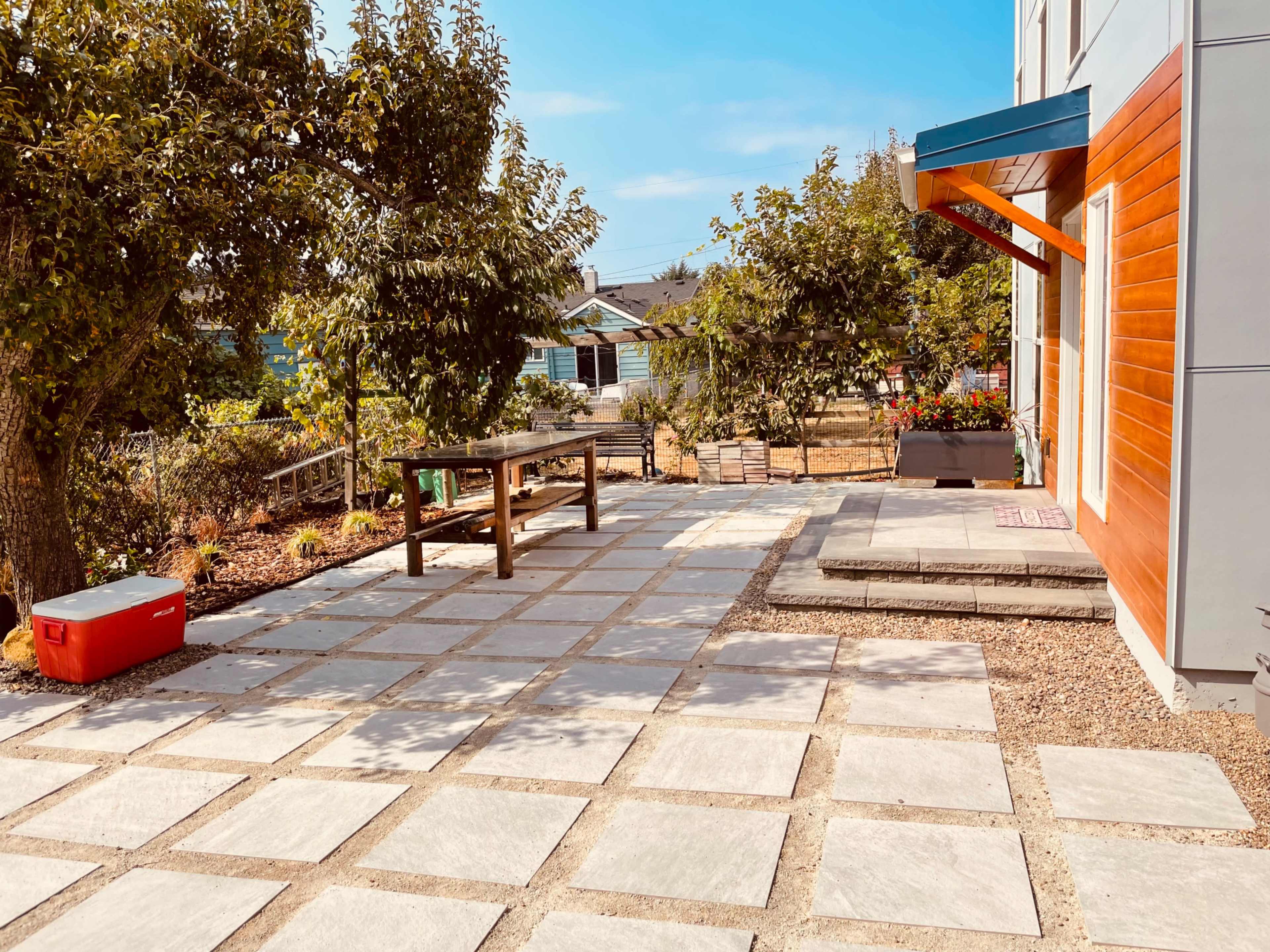A landscaped outdoor area features a stone patio with a wooden table, surrounded by trees and gardens.