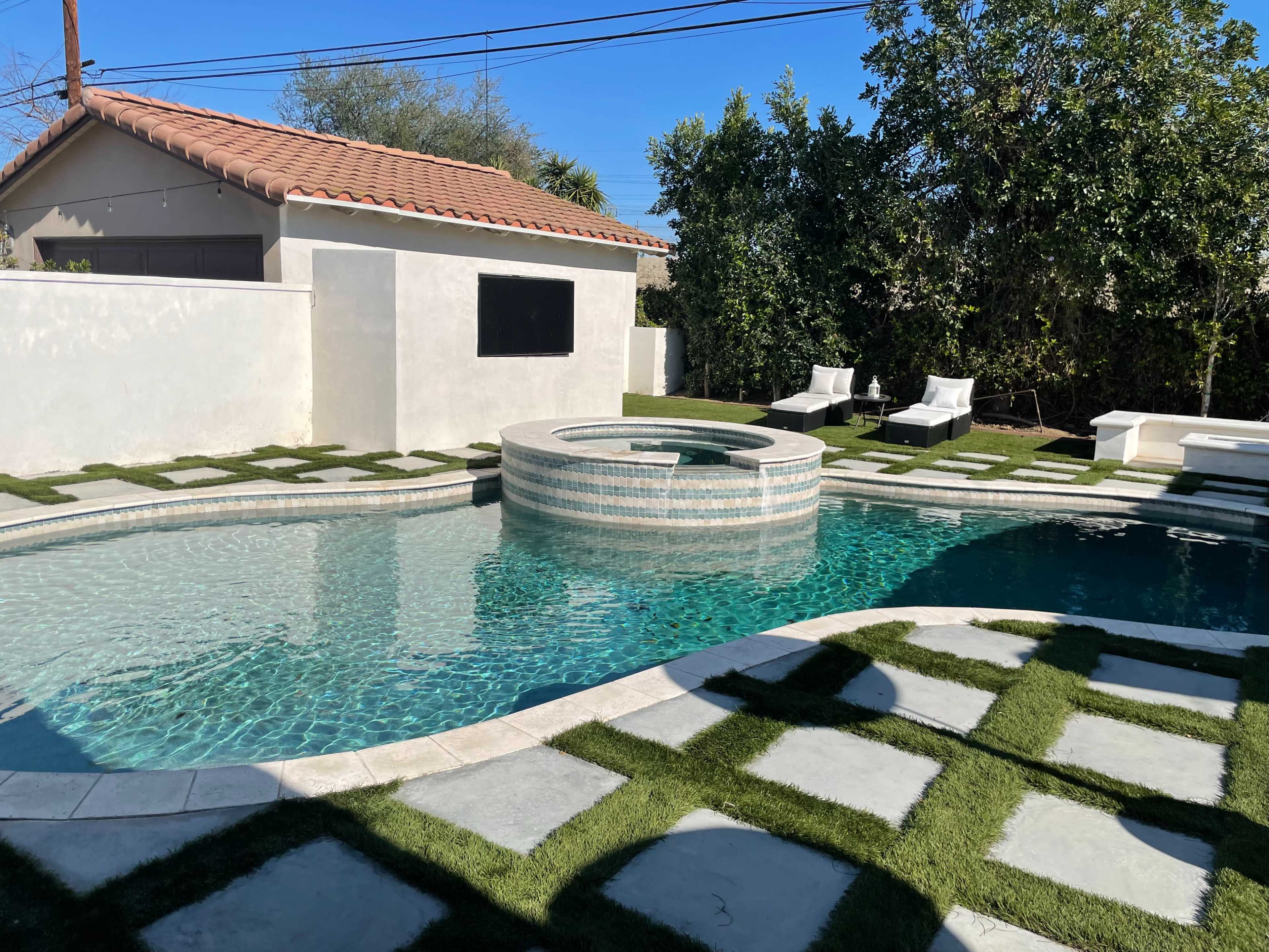 A landscaped backyard featuring a swimming pool with a spa, surrounded by artificial grass and stone pathways, along with lounge chairs positioned nearby.