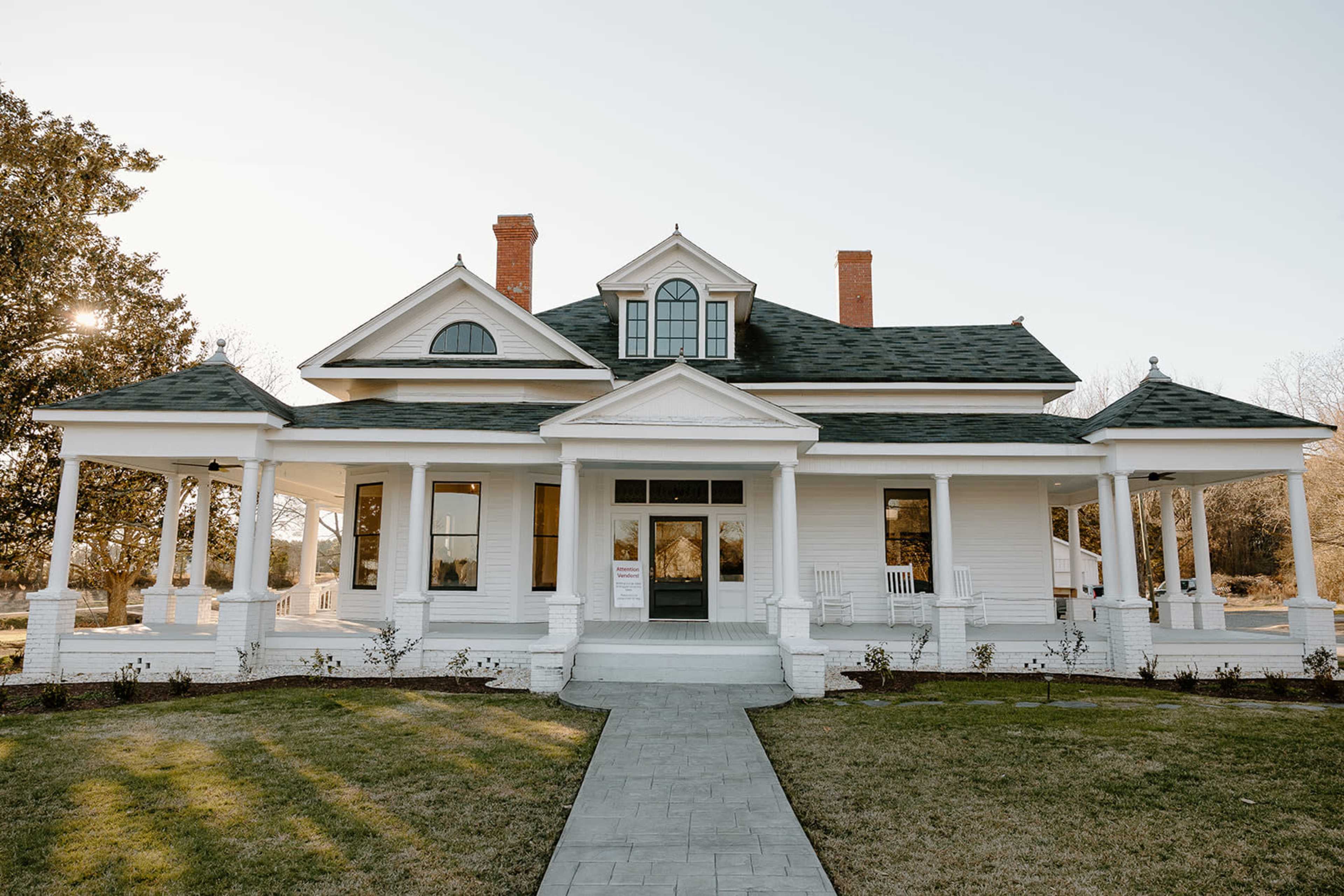 A large white house with a green roof features a central entrance flanked by two porches and surrounded by a well-manicured lawn.