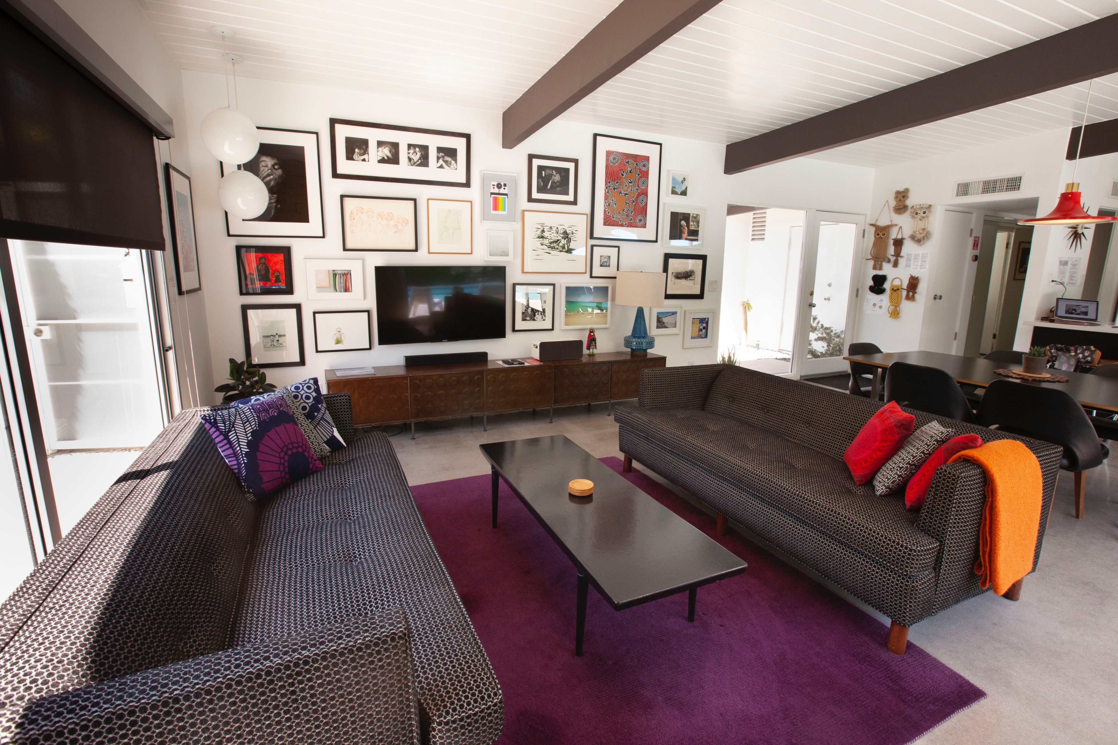 The image shows a modern living room with two patterned sofas facing a low coffee table, a wall adorned with framed artworks, and a large television on a wooden console.