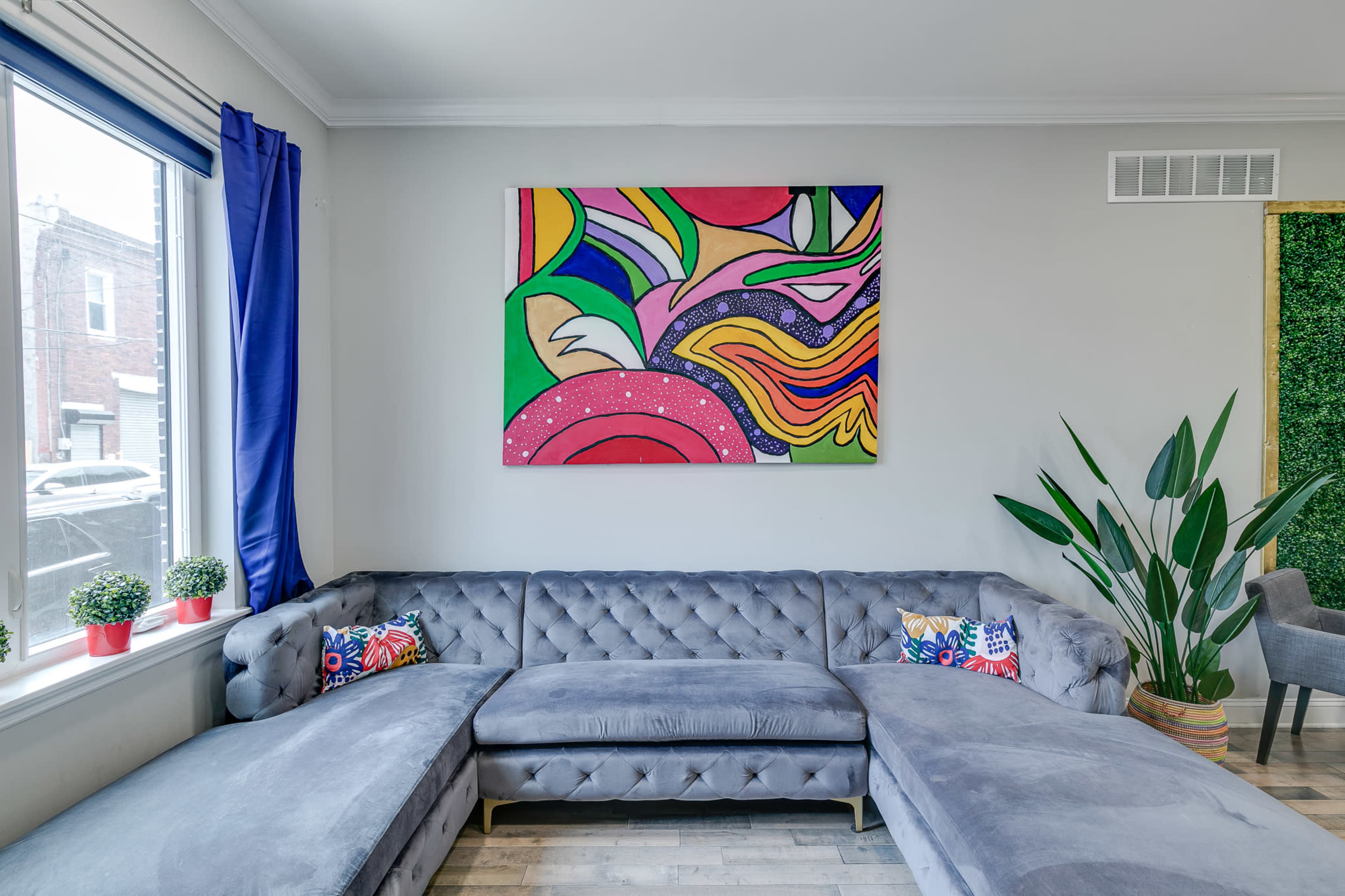 A modern living room features a gray sectional sofa, colorful artwork on the wall, and potted plants by the window.