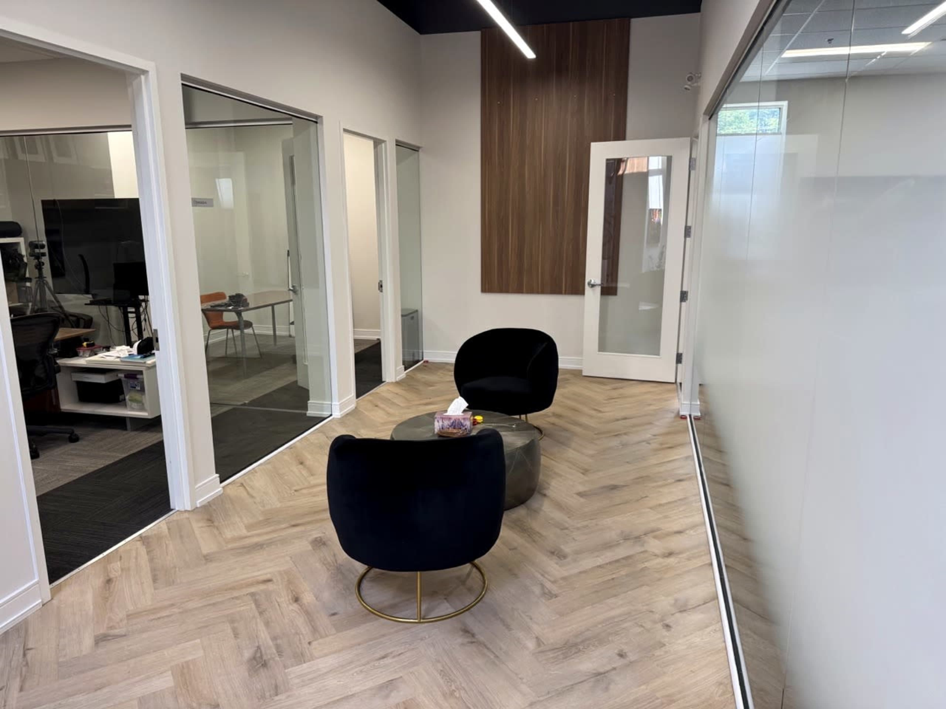 The image shows a modern office corridor with two chairs and a small table in the center, flanked by glass-walled rooms on one side and a wooden panel wall on the other.