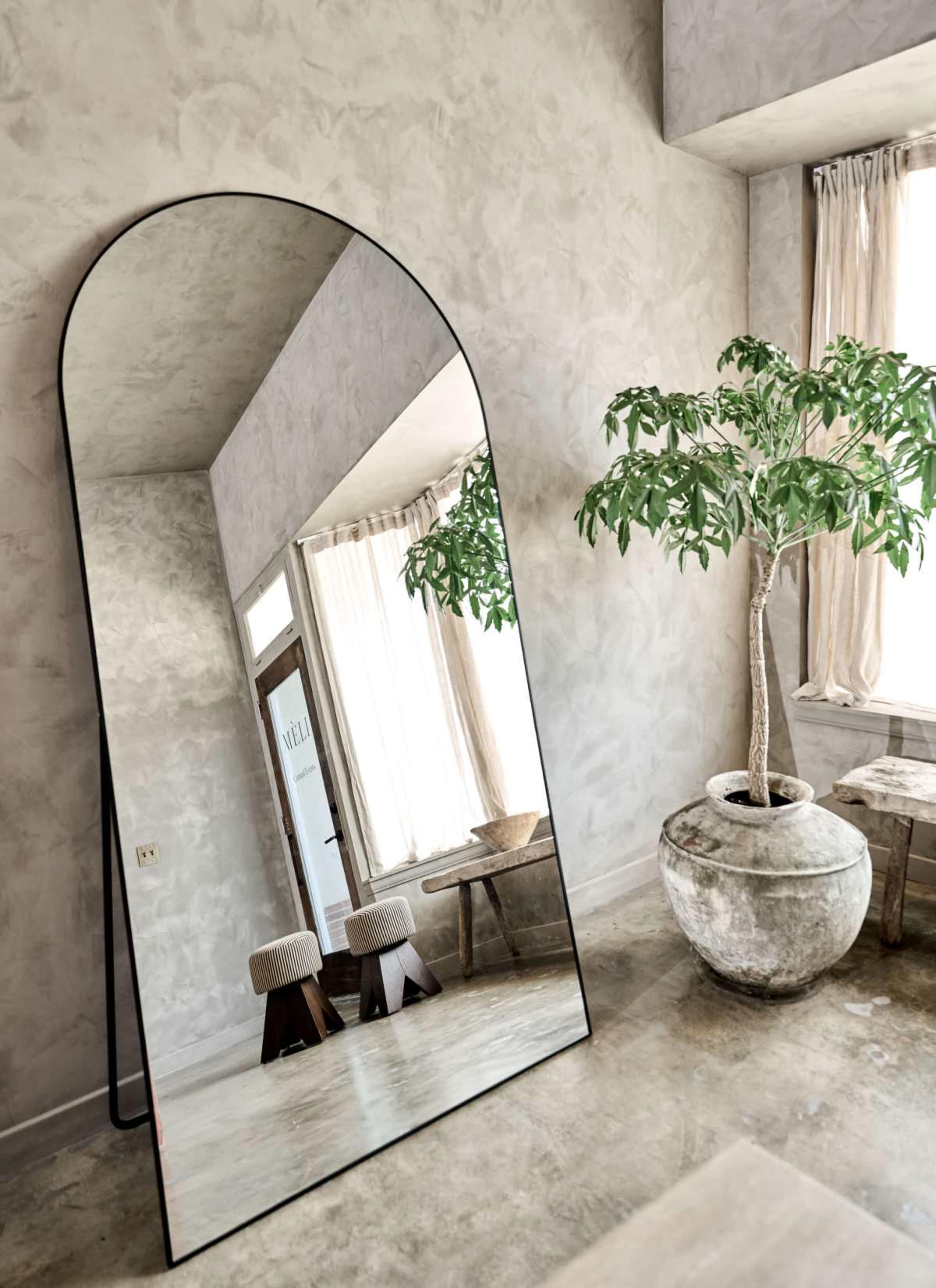 A large, arched mirror leans against a wall in a room with minimalistic decor, featuring a potted plant, a wooden table, and natural light streaming through sheer curtains.