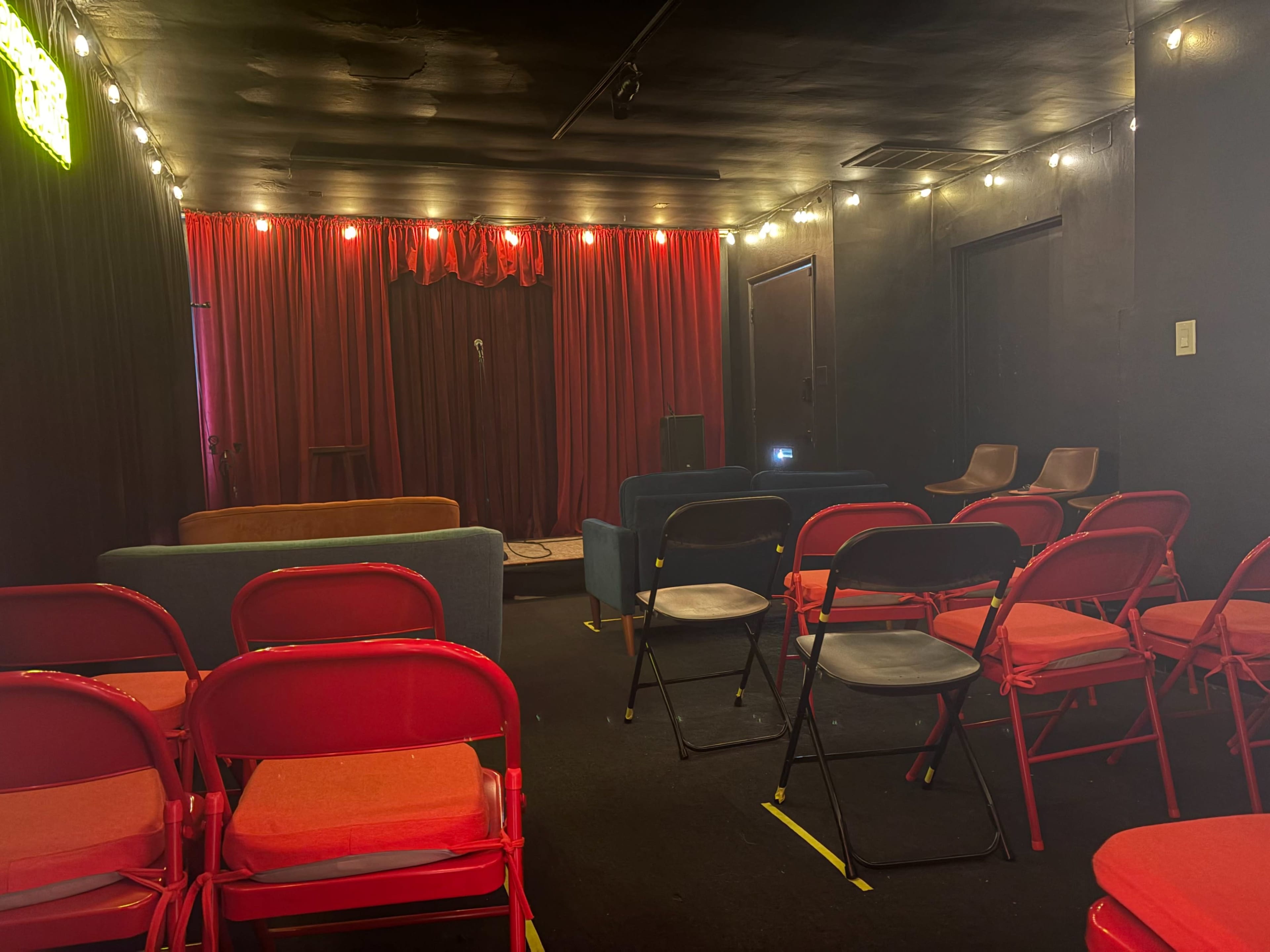 The image shows a small performance space with a stage, red curtains, and seating arranged in rows, including red and black chairs, along with a few sofas at the back.