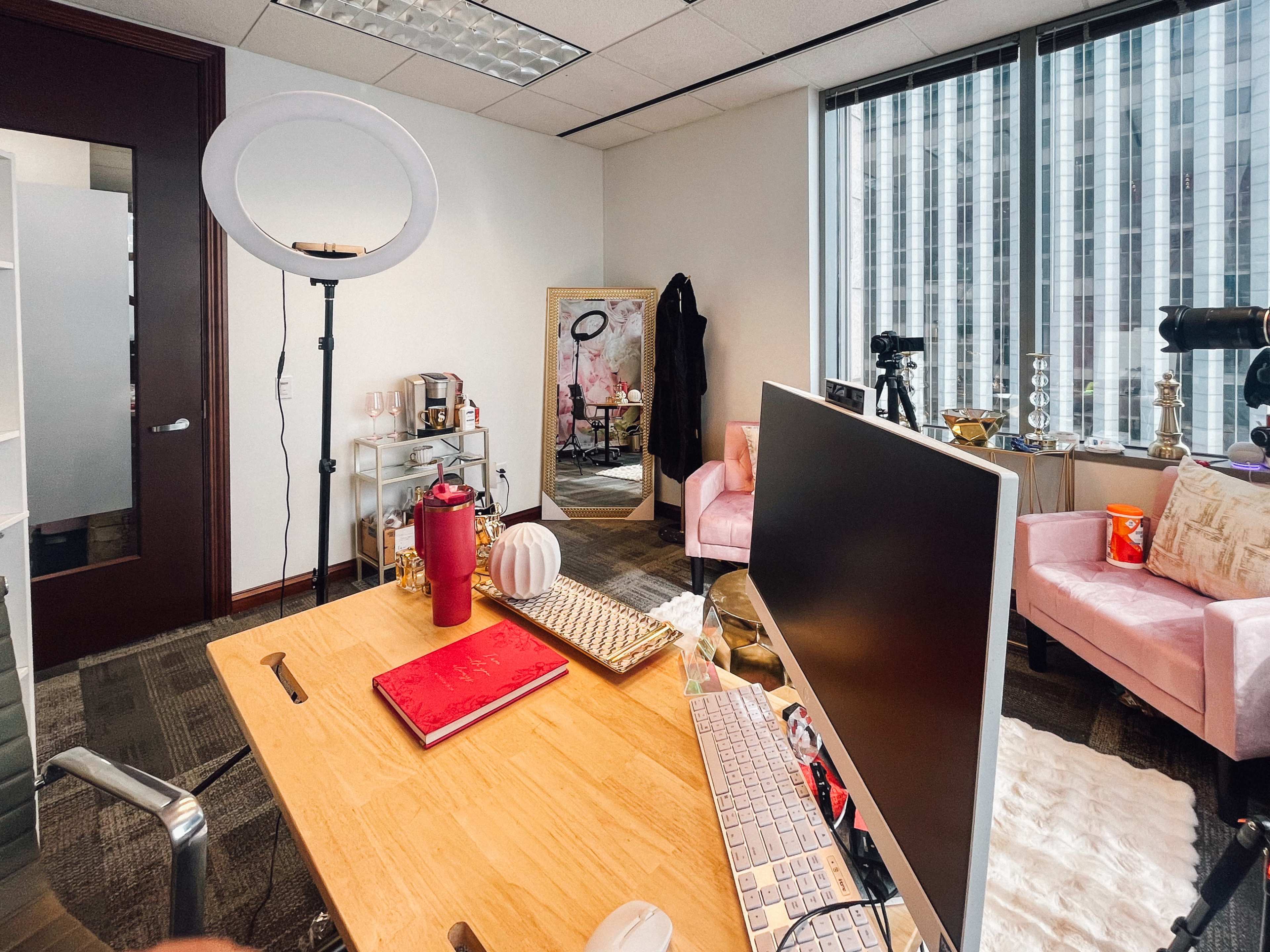 The image shows a well-lit office space featuring a wooden desk with a computer, a ring light, pink chairs, and large windows with a city view.