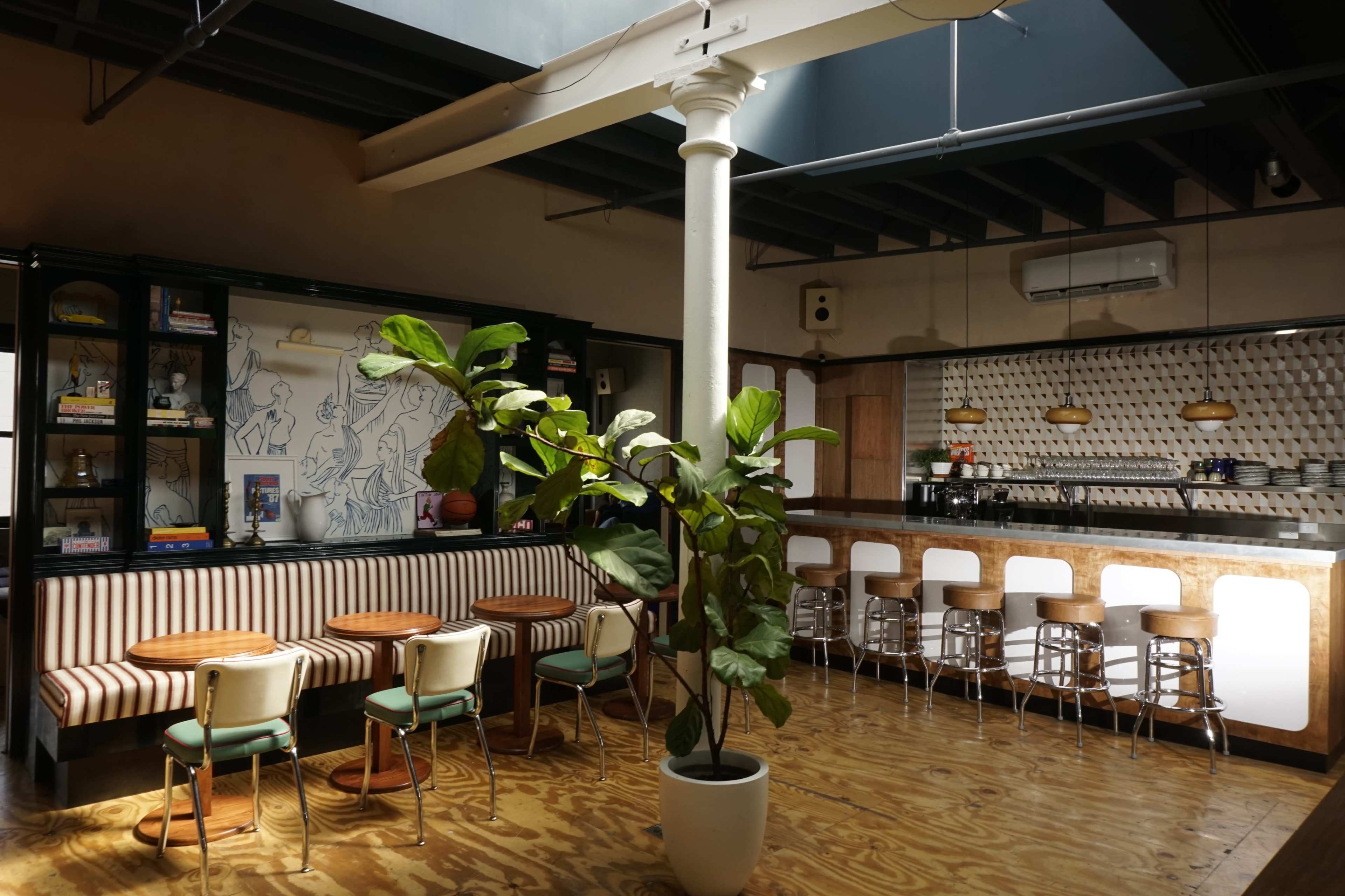 The image shows a modern café interior with a central column, a large plant, striped seating, and a bar area with stools.