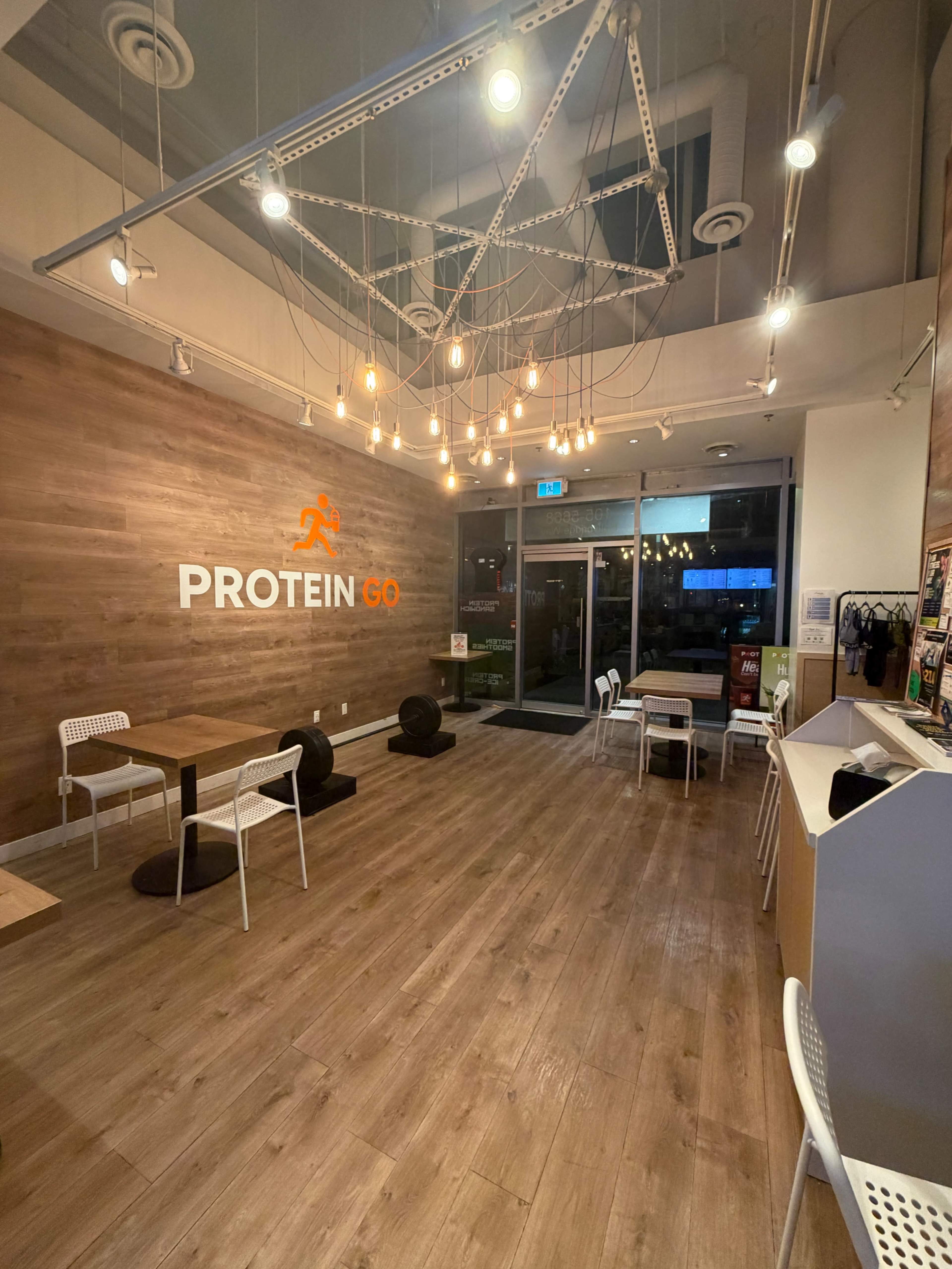 The interior of a cafe named "Protein Go" features wooden walls, modern lighting, and simple white furniture.
