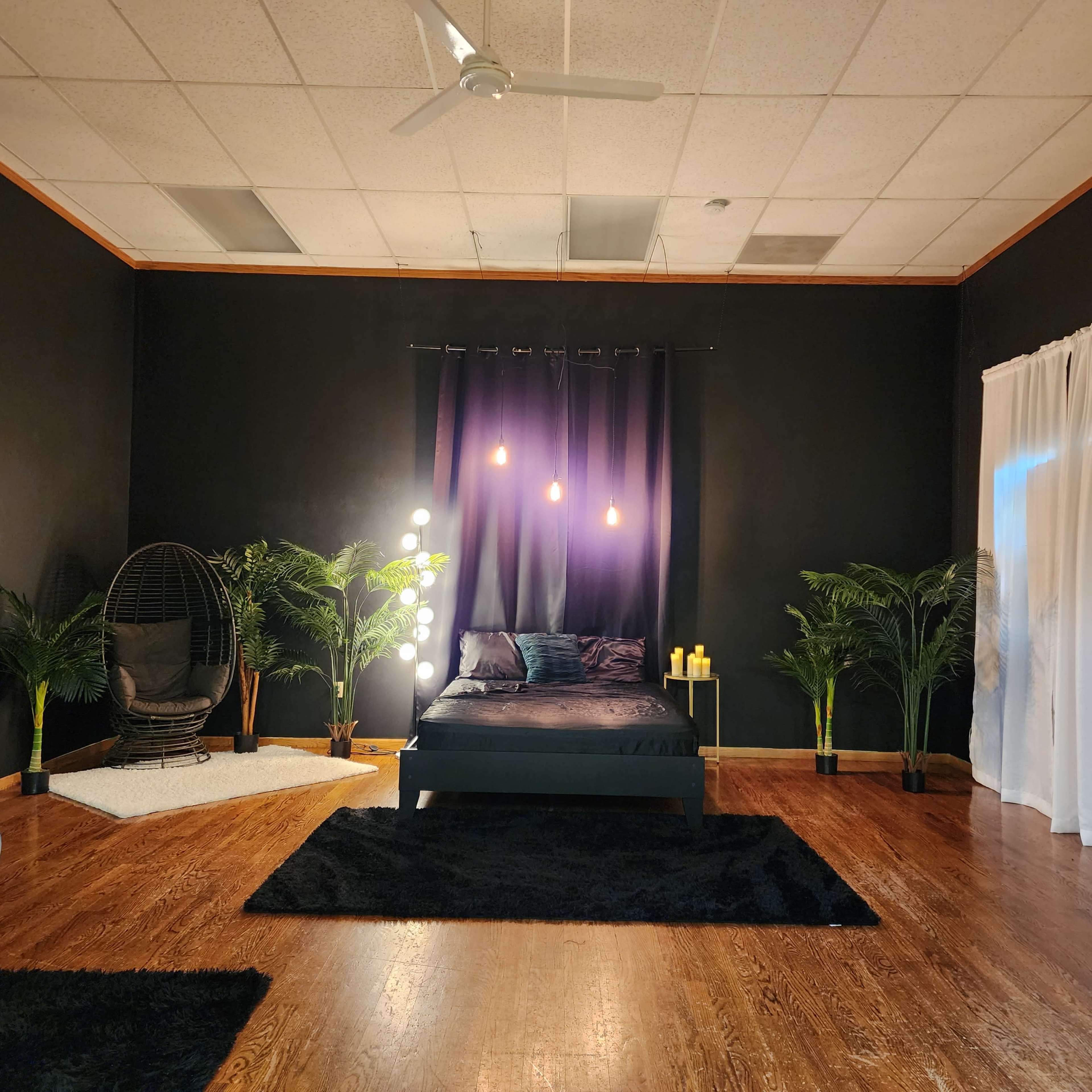 A dark-themed bedroom with a black bed, hanging lights, and various indoor plants, complemented by a hanging chair and rugs on the wooden floor.