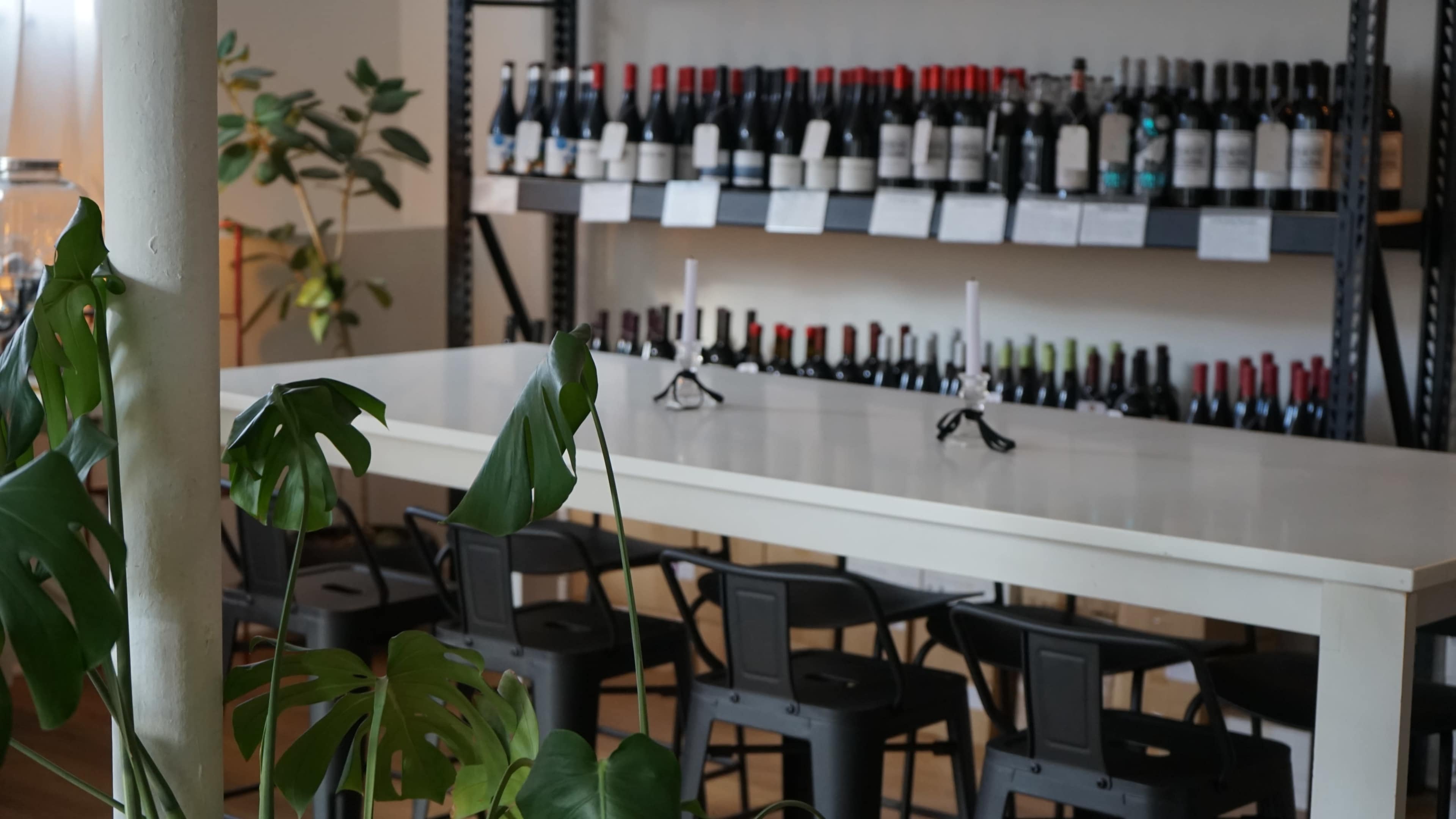 A spacious wine bar features a long white table with black chairs, lined with a collection of wine bottles on shelves in the background.