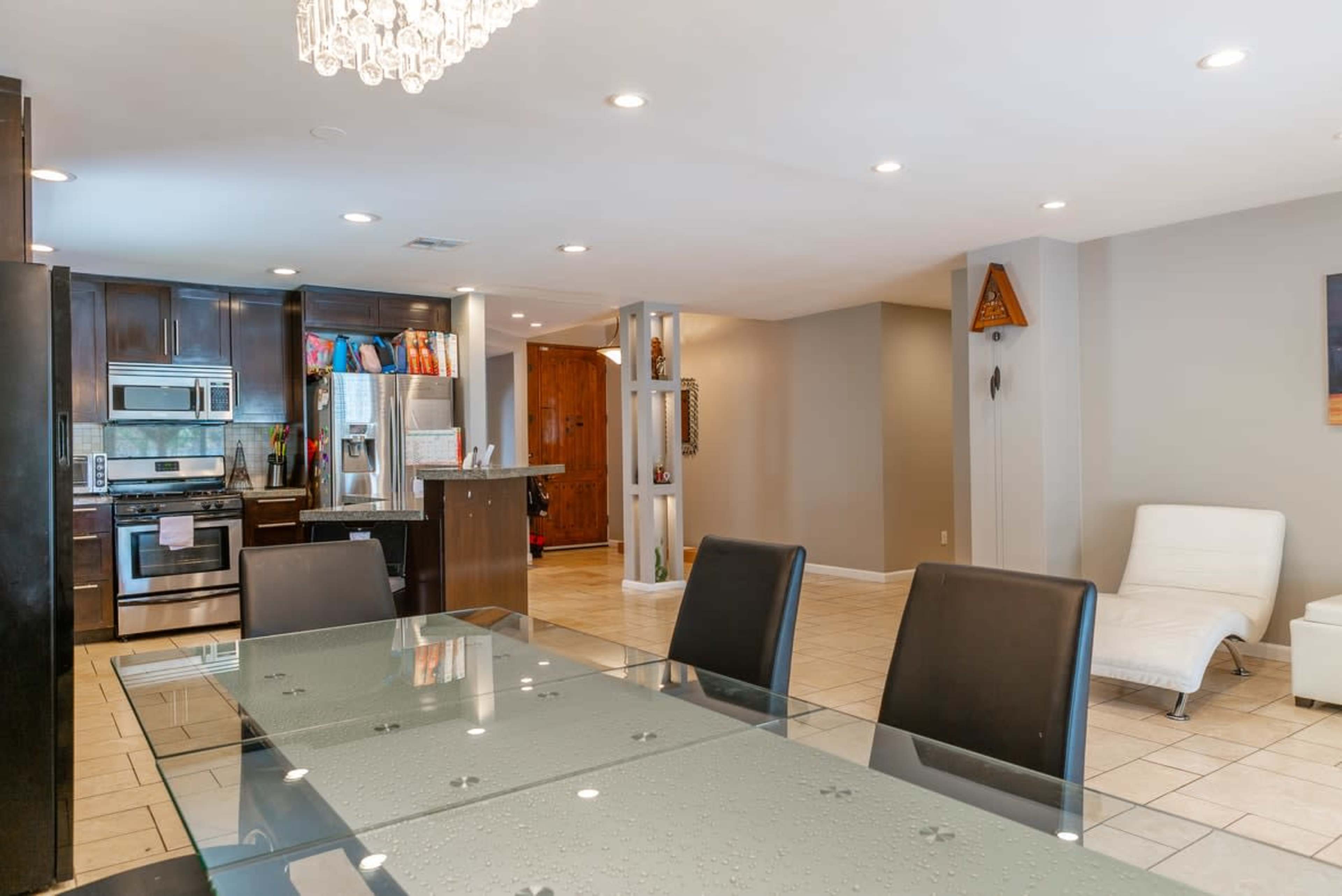 The image shows a modern dining area with a glass table and chairs, leading into a kitchen featuring dark cabinetry and stainless steel appliances.