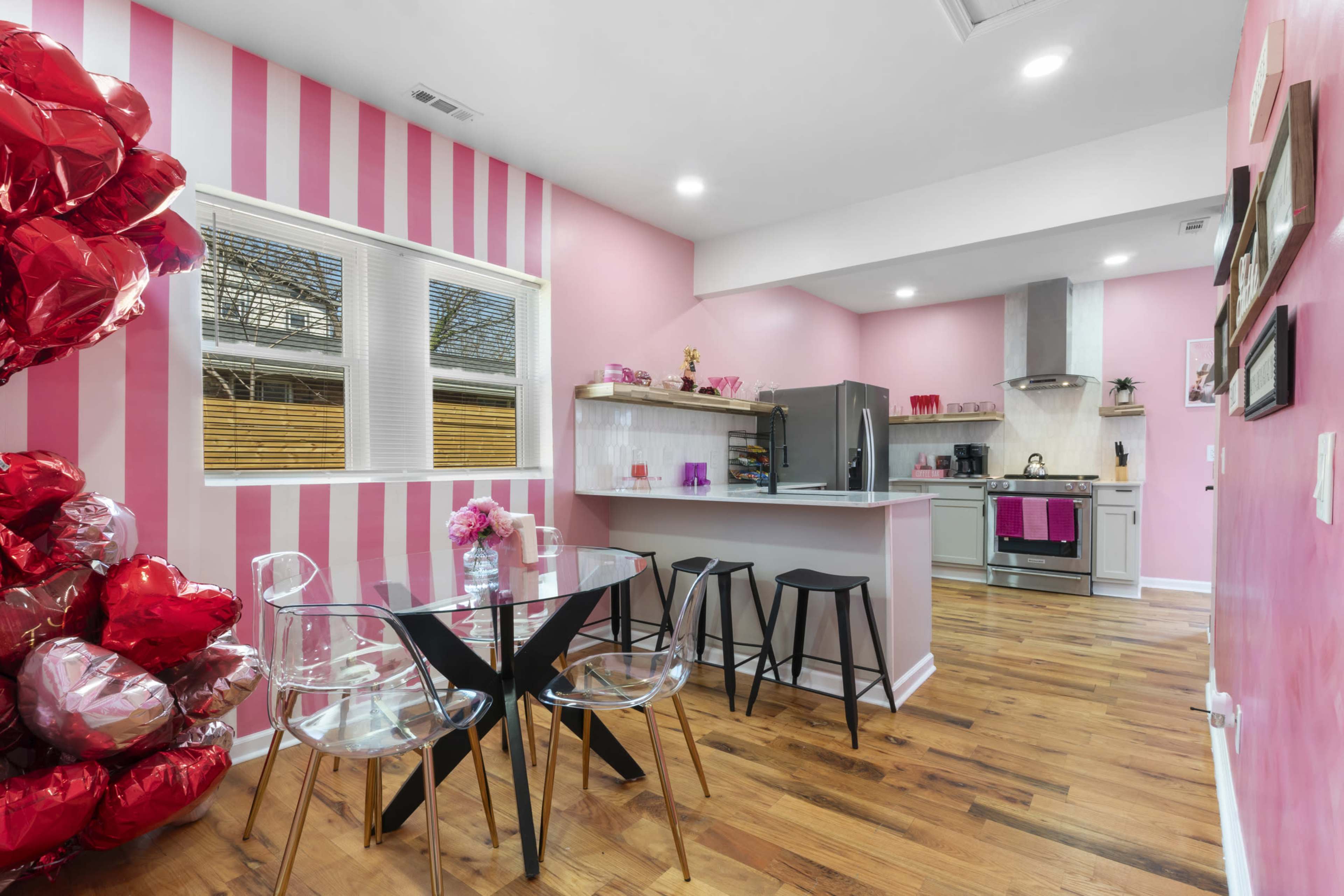 The image shows a modern kitchen and dining area with pink-striped walls, a glass dining table, and a cluster of red heart-shaped balloons nearby.