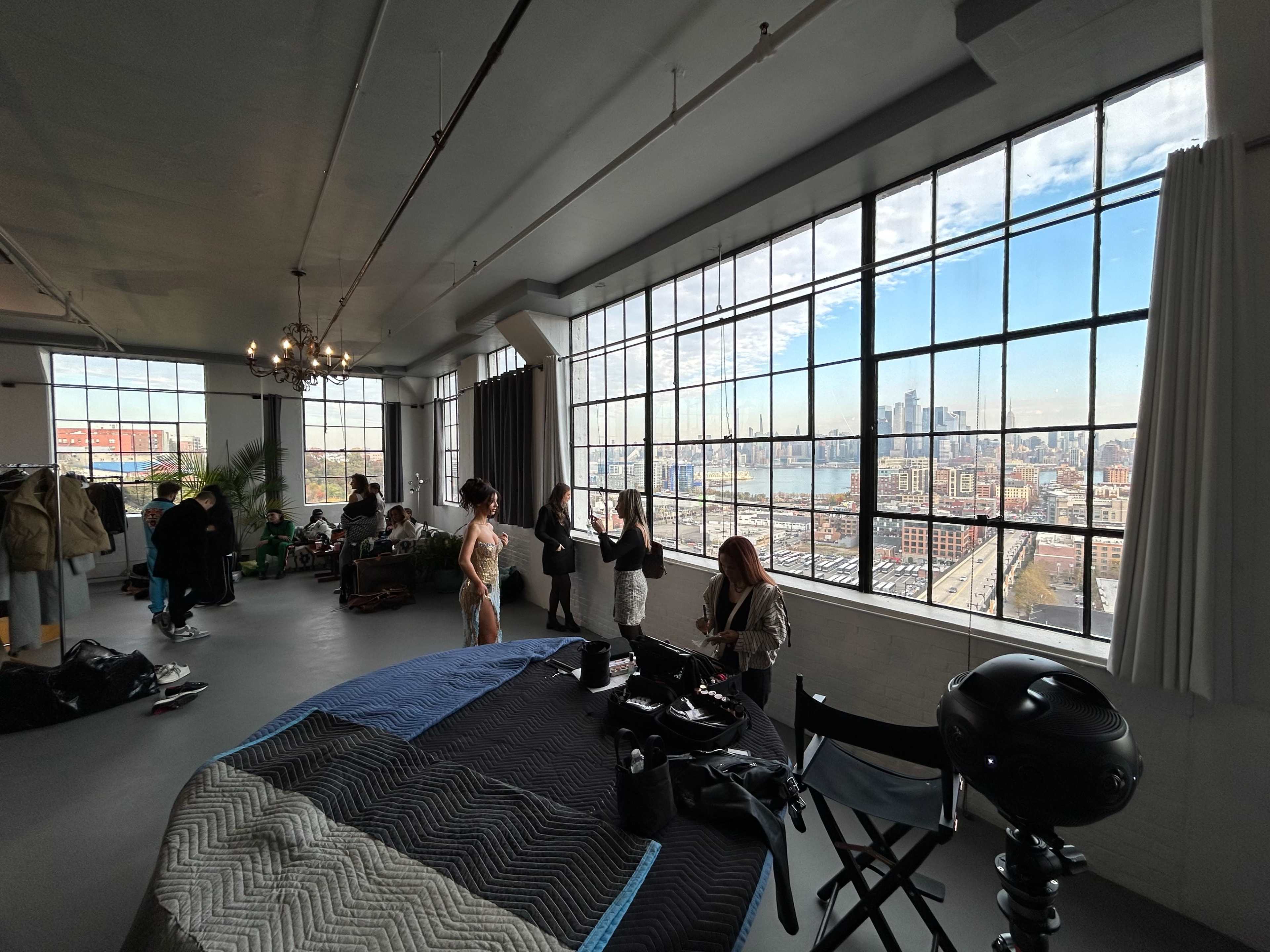 A bright studio with large windows overlooks an urban skyline, while people are preparing and organizing equipment inside.