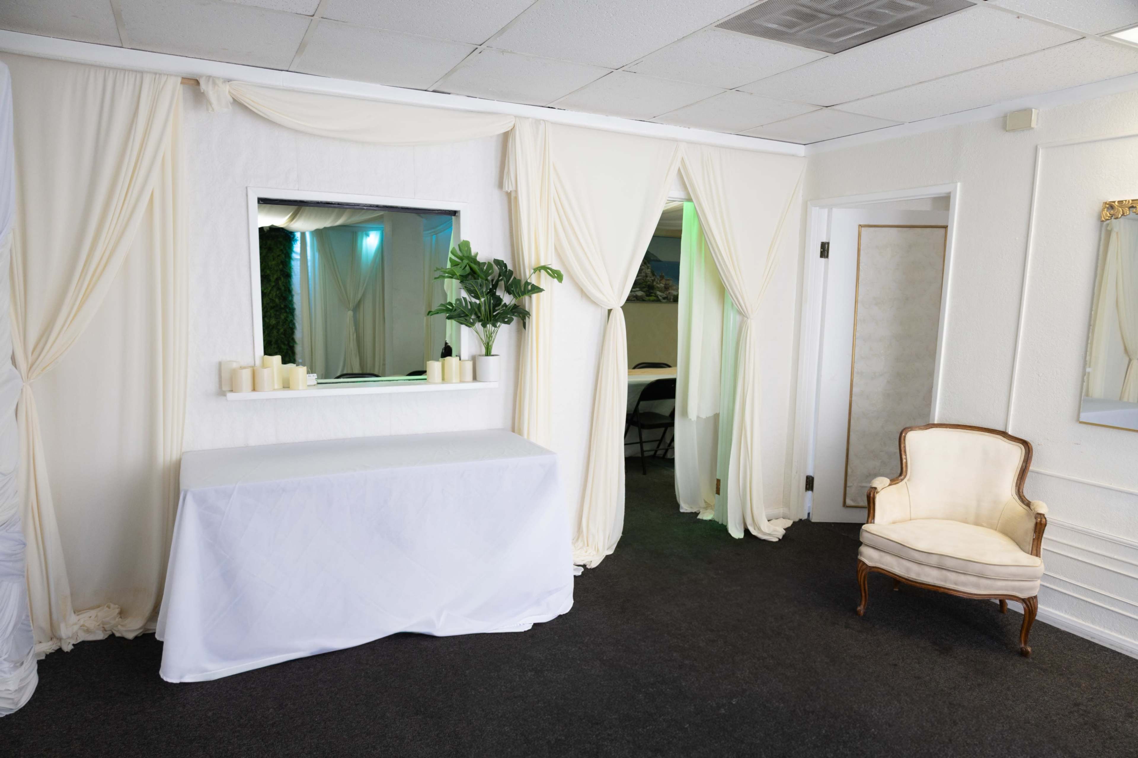 The image shows a room featuring white drapery, a table covered with a white cloth, a framed mirror on the wall, a green plant, and a vintage chair near the doorway.