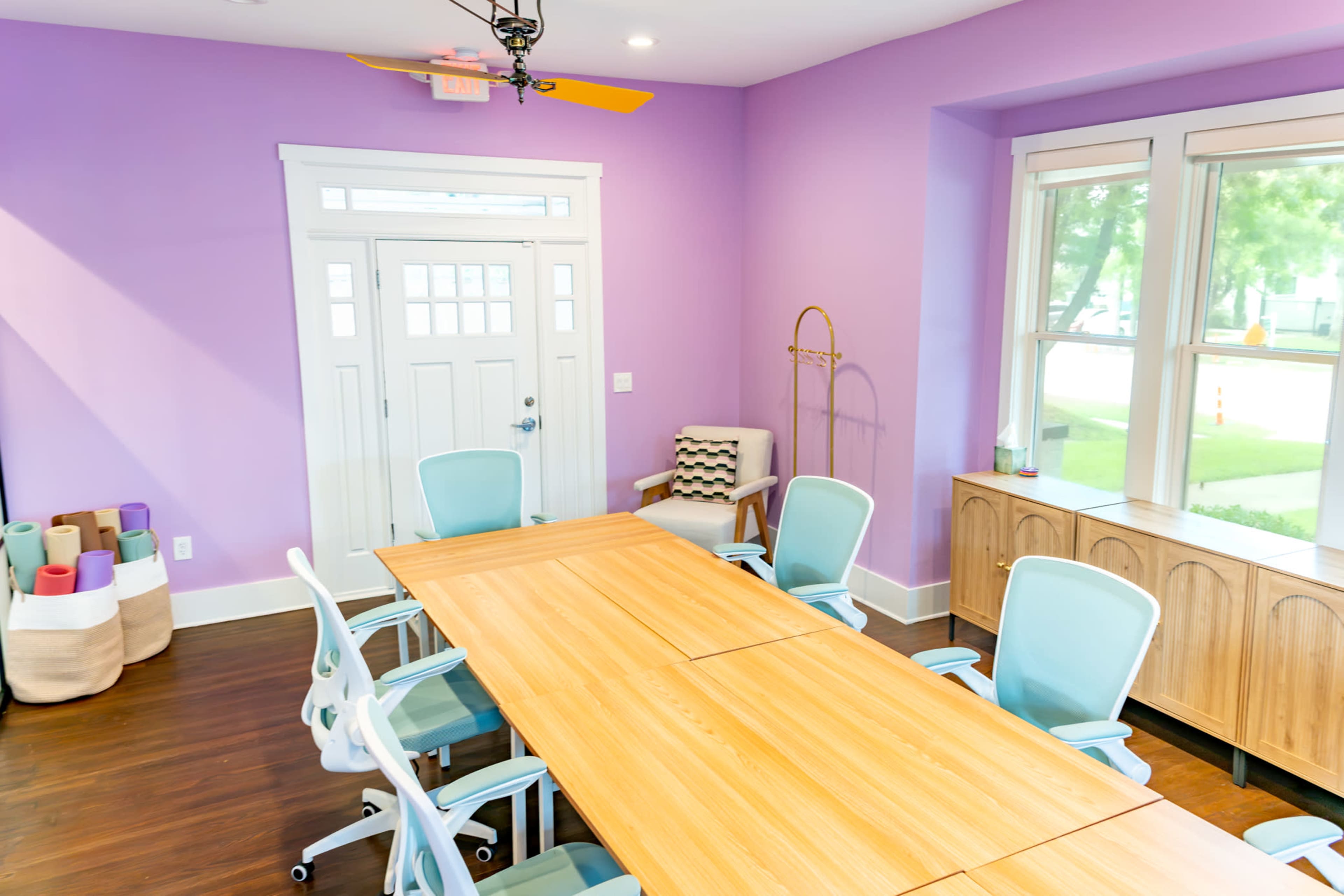 A conference room features a large wooden table surrounded by light blue chairs, with purple walls and a white door leading outside.