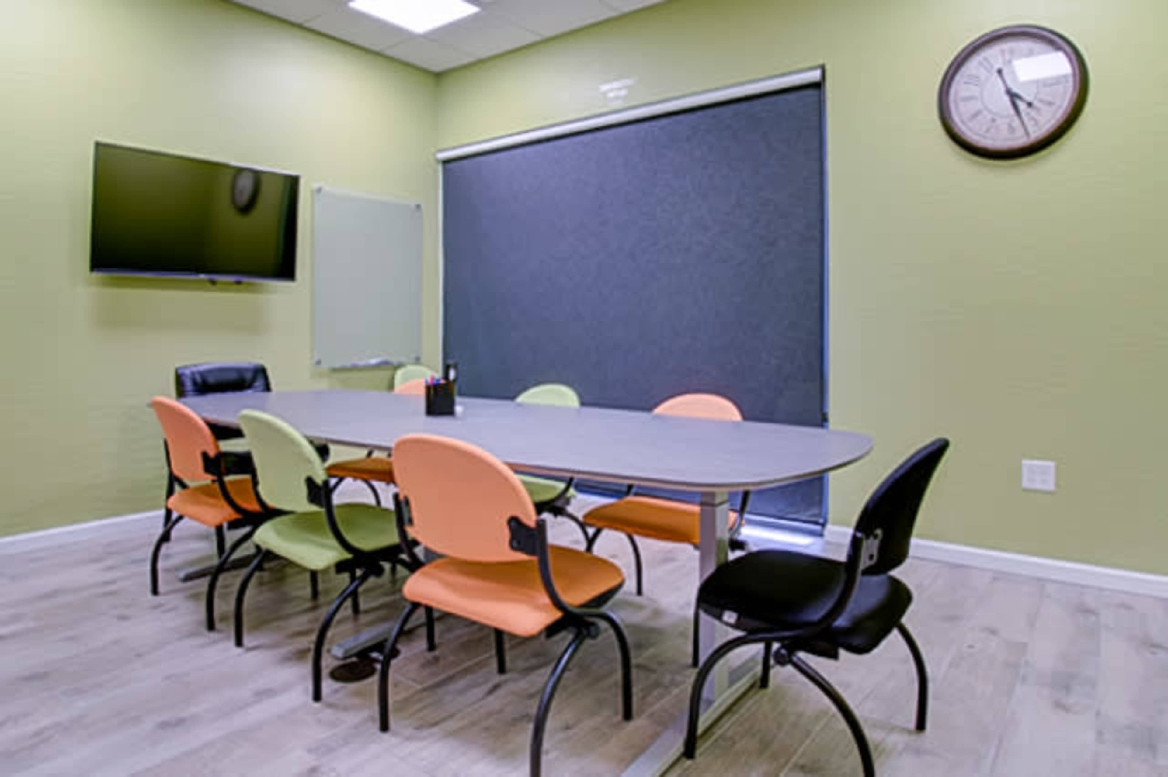 A meeting room features a large table surrounded by chairs in orange and black, with a TV mounted on the wall and a clock above the door.
