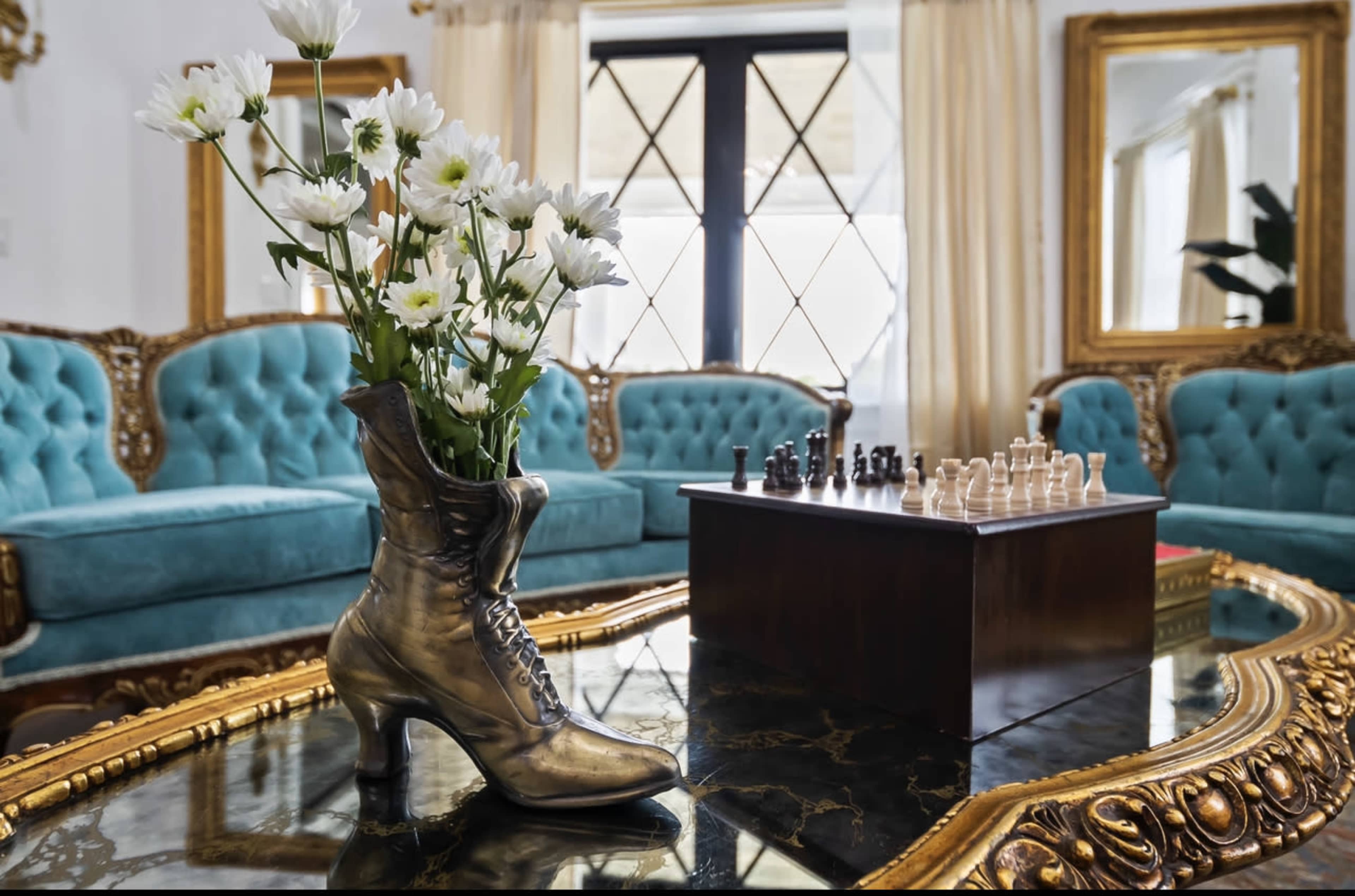 A vintage boot holds white daisies on a coffee table in a living room featuring tufted turquoise sofas and a chess set.