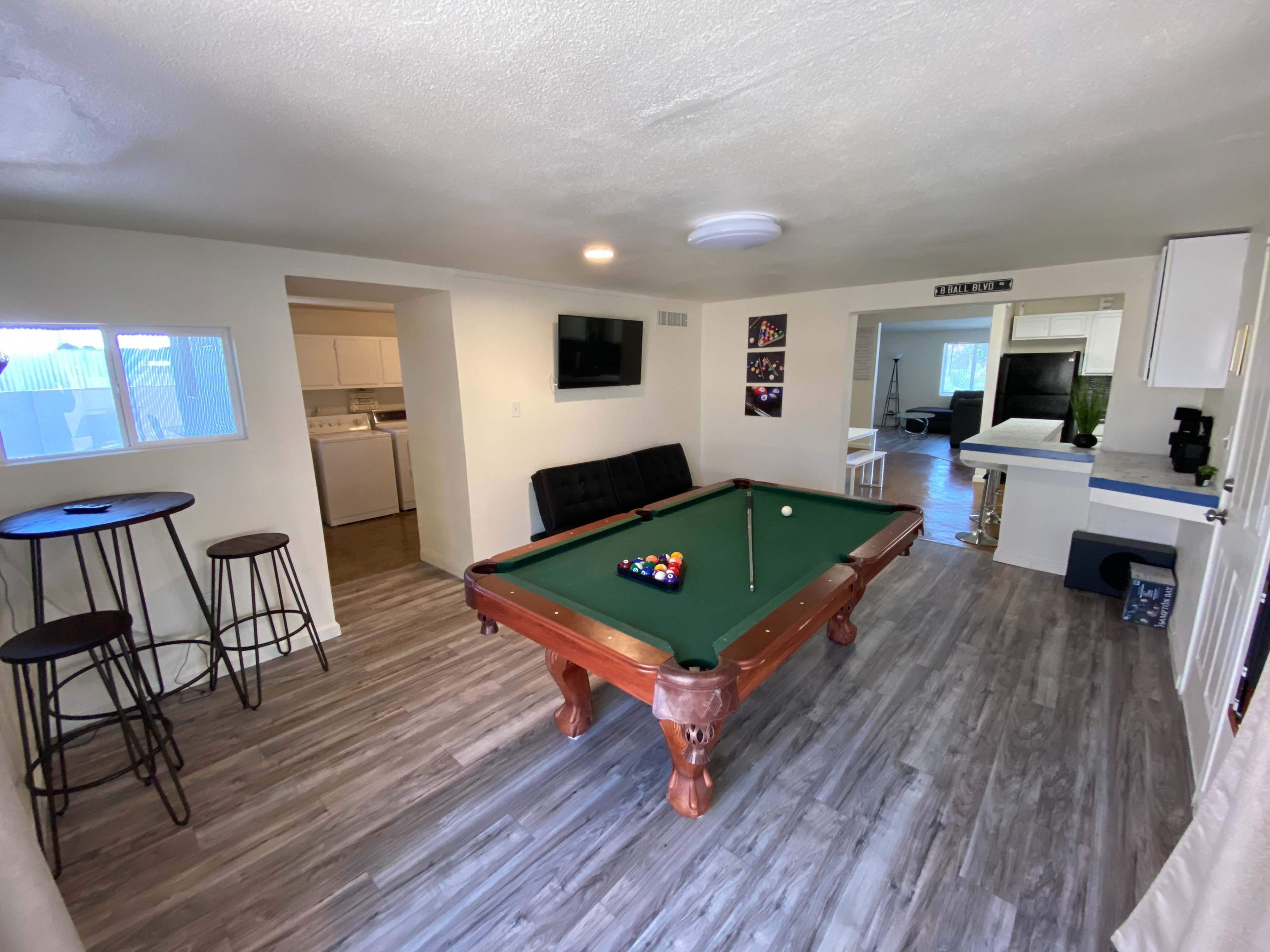 A room featuring a pool table, bar stools, and a kitchenette visible through an open doorway.