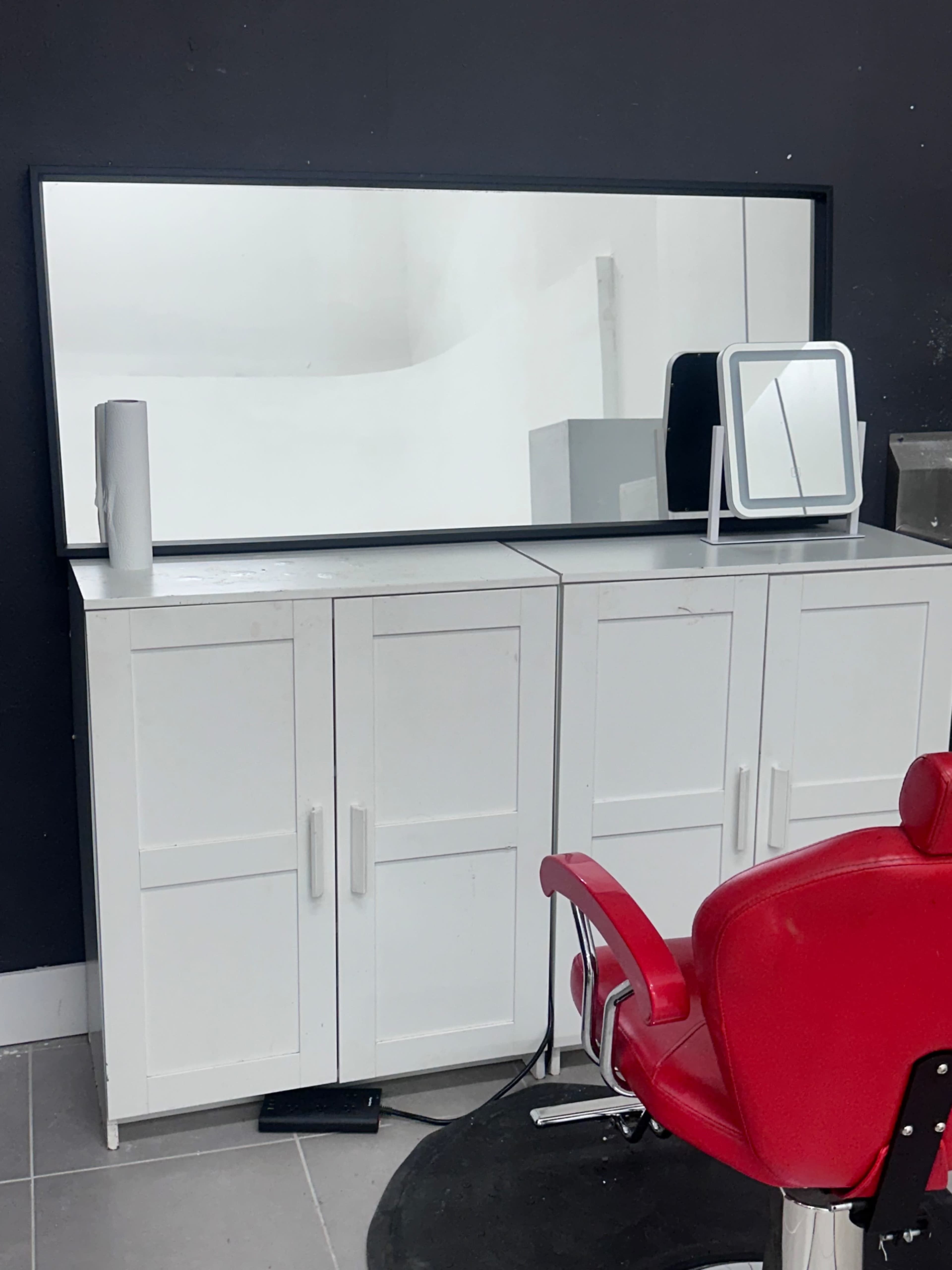 A white cabinet with two doors is positioned underneath a large rectangular mirror in a room featuring a red barber chair.