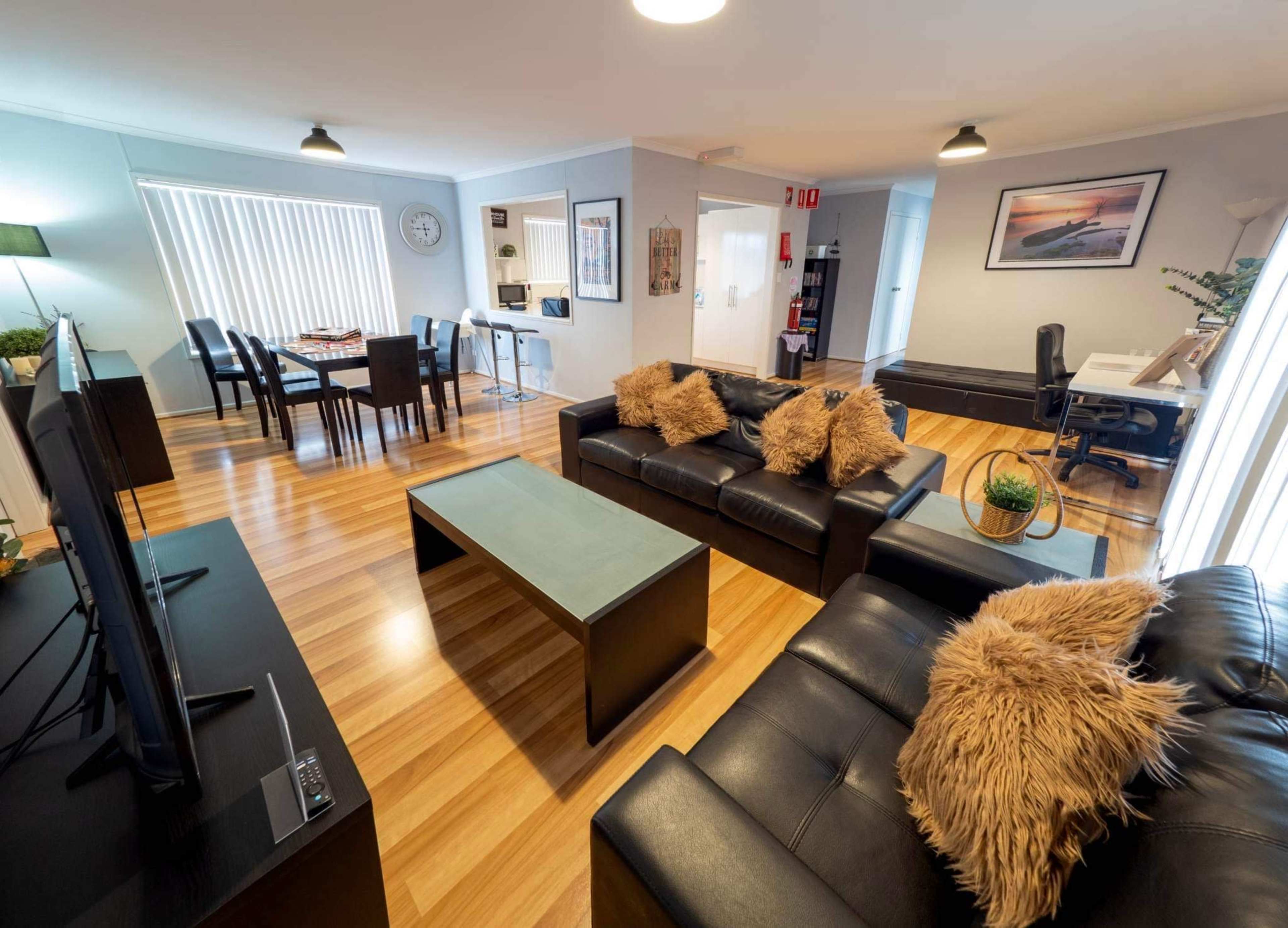The image shows a modern living room with black leather couches, a coffee table, and wooden flooring, adjacent to a dining area with a table and chairs.