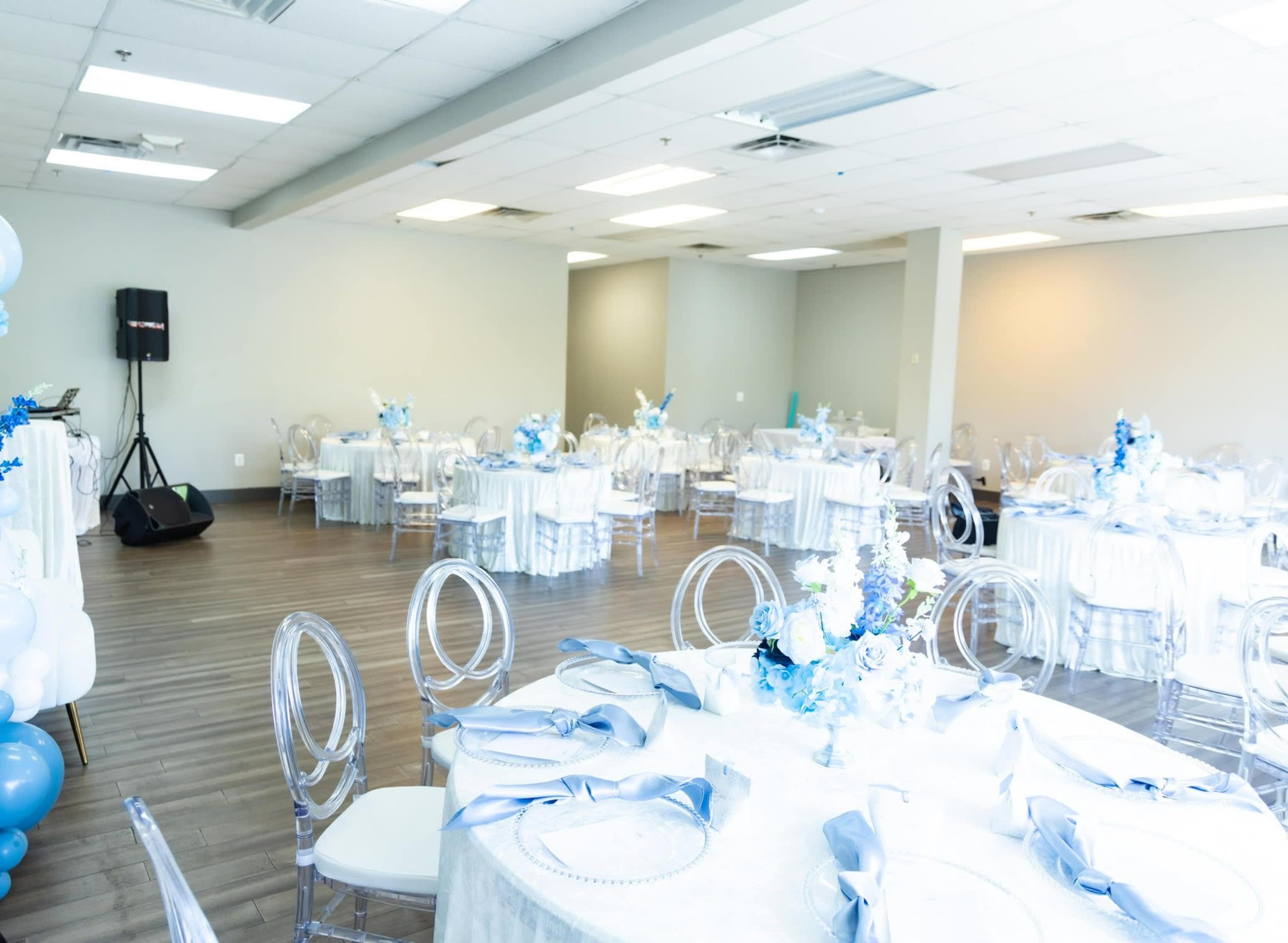 The image shows a spacious event room arranged with elegant round tables, each covered with white tablecloths and decorated in shades of blue, accompanied by transparent chairs.