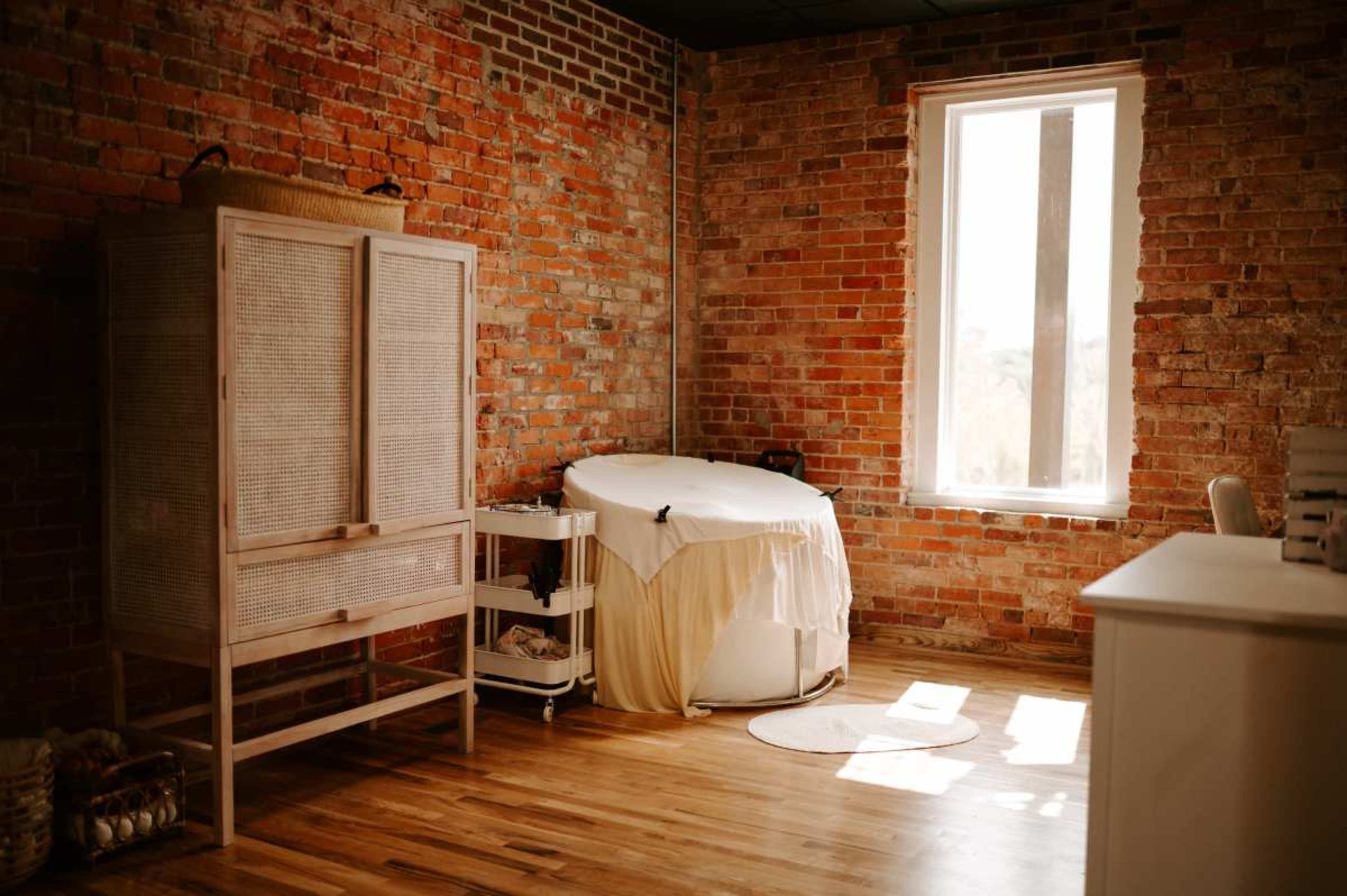 A cozy room with exposed brick walls, featuring a massage table draped in white linens, a wooden cabinet, and a large window allowing natural light to enter.