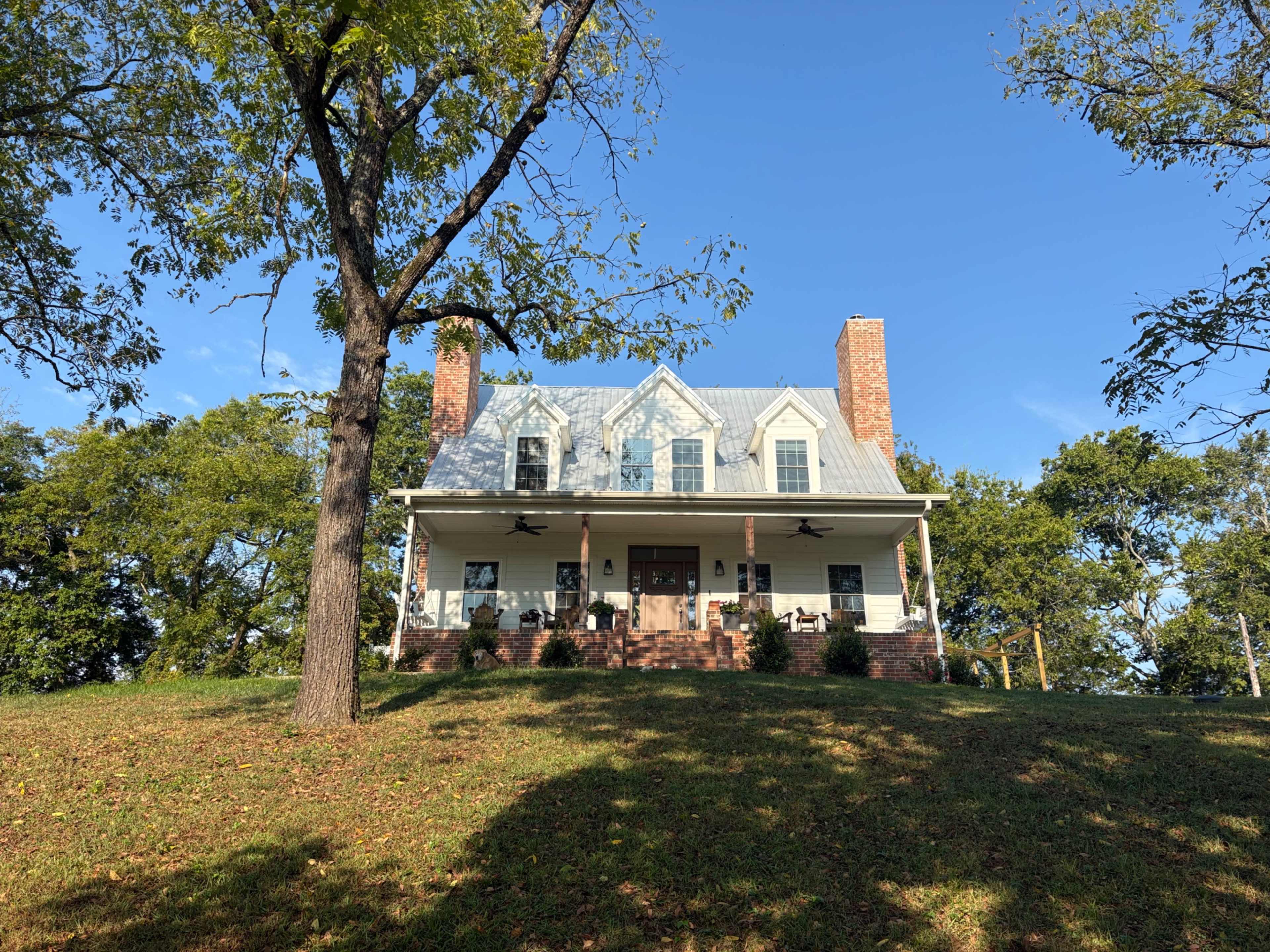 A two-story house with a gabled roof, porch, and brick chimneys sits atop a grassy hill surrounded by trees under a clear blue sky.