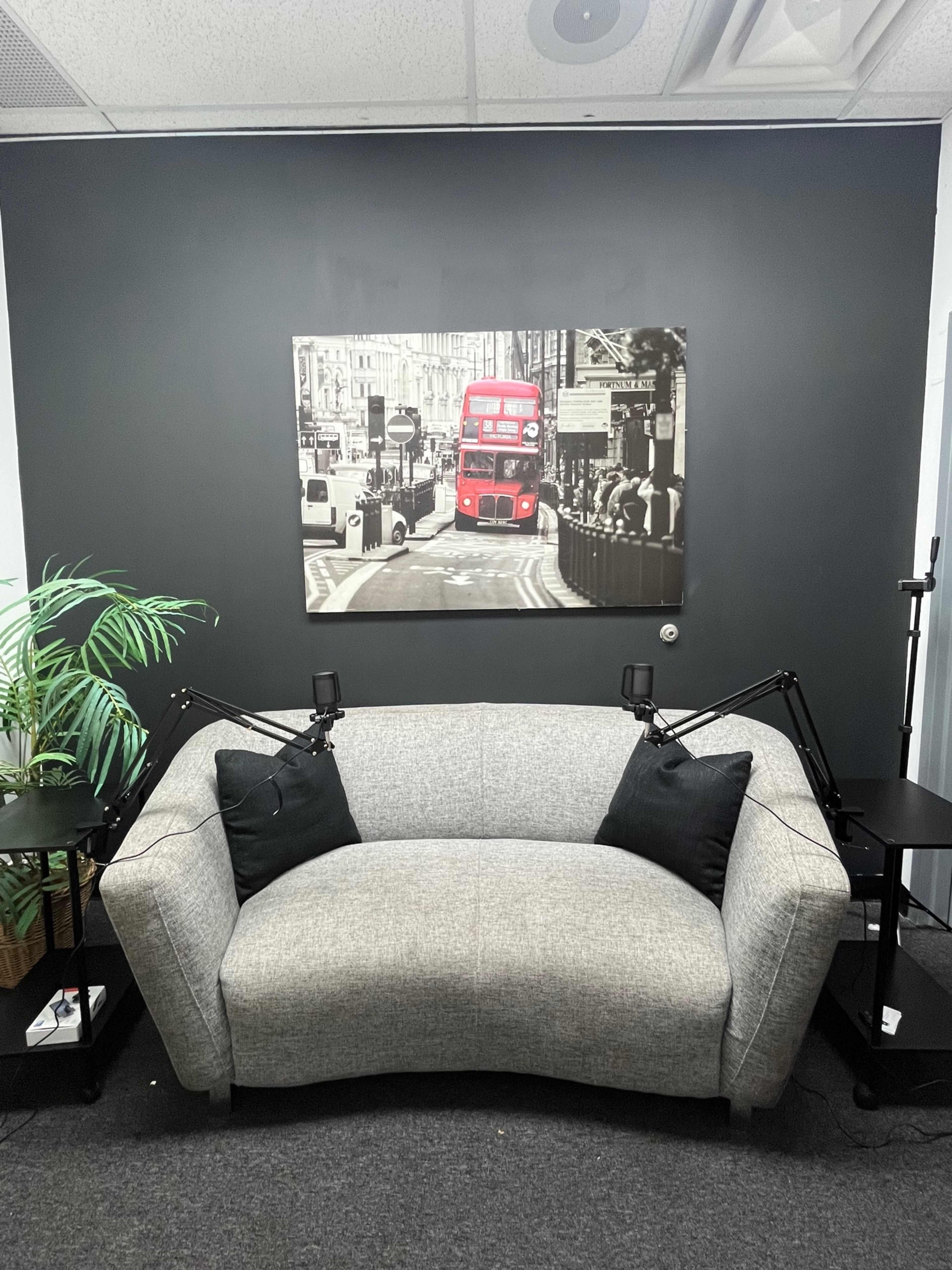 A gray couch with black cushions is positioned against a dark wall, featuring a black-and-white photograph of a red double-decker bus in an urban setting above it, with two black side tables and a green plant nearby.