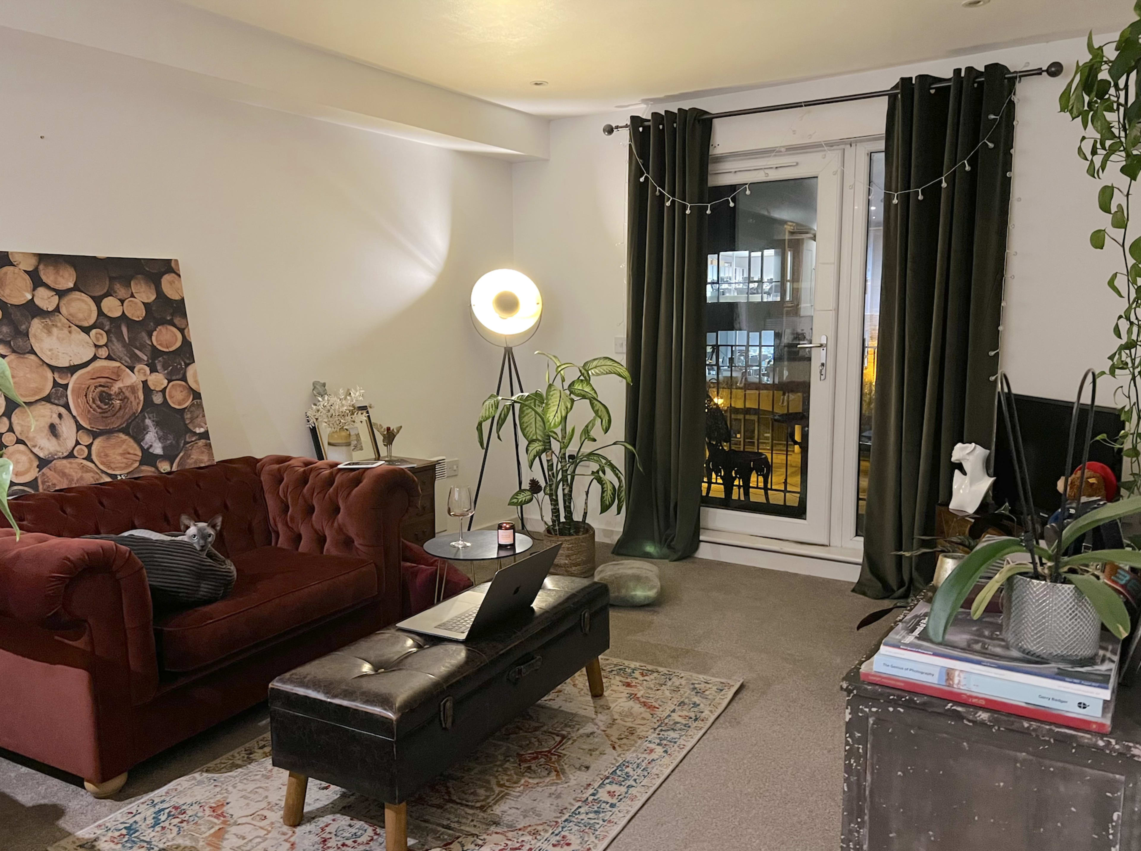 A cozy living room features a red tufted sofa, a large floor lamp, and a view of a balcony with green plants and decorative elements.