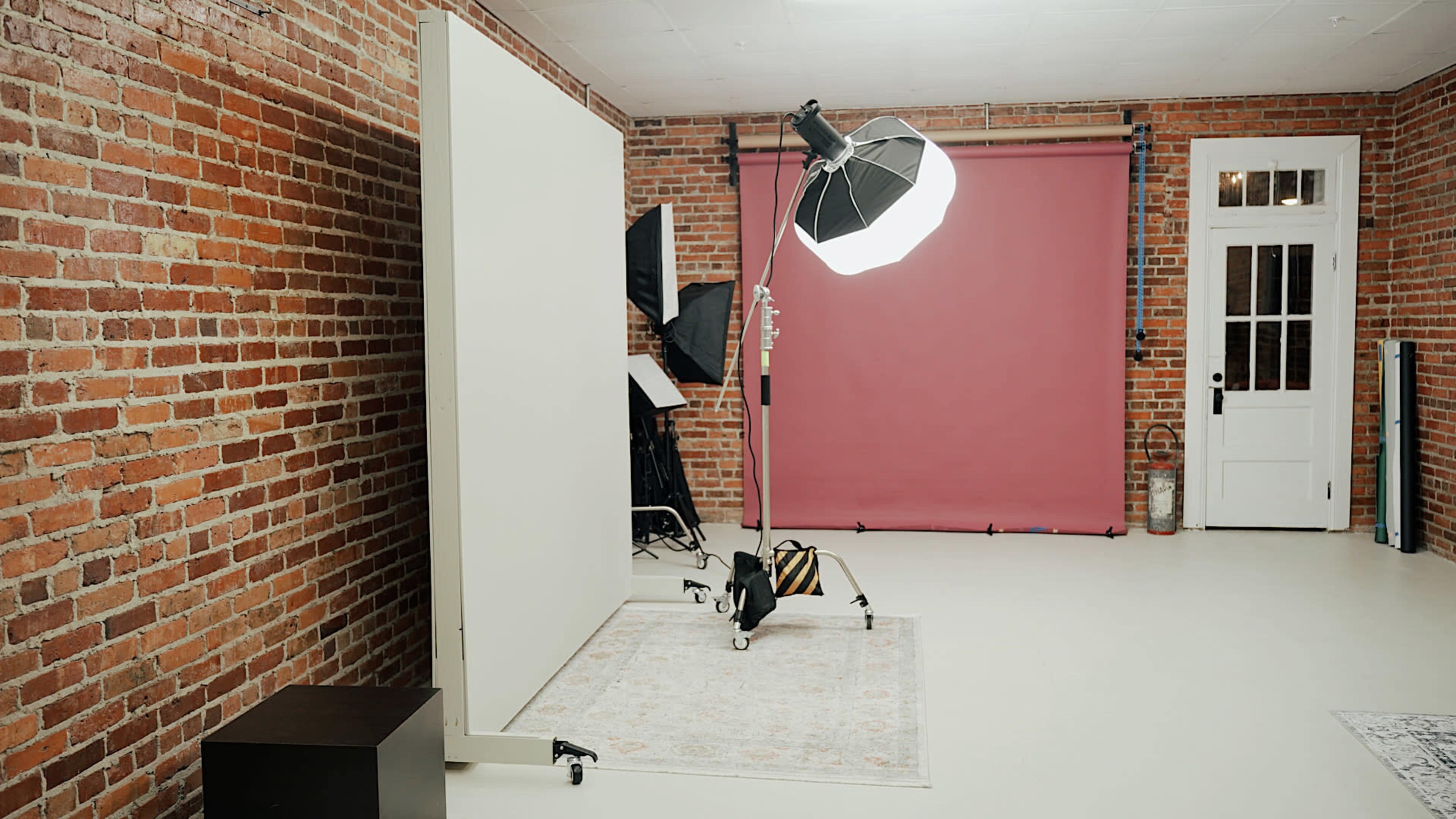 The image shows a photography studio with a brick wall, a light setup, a pink backdrop, and a white partition.
