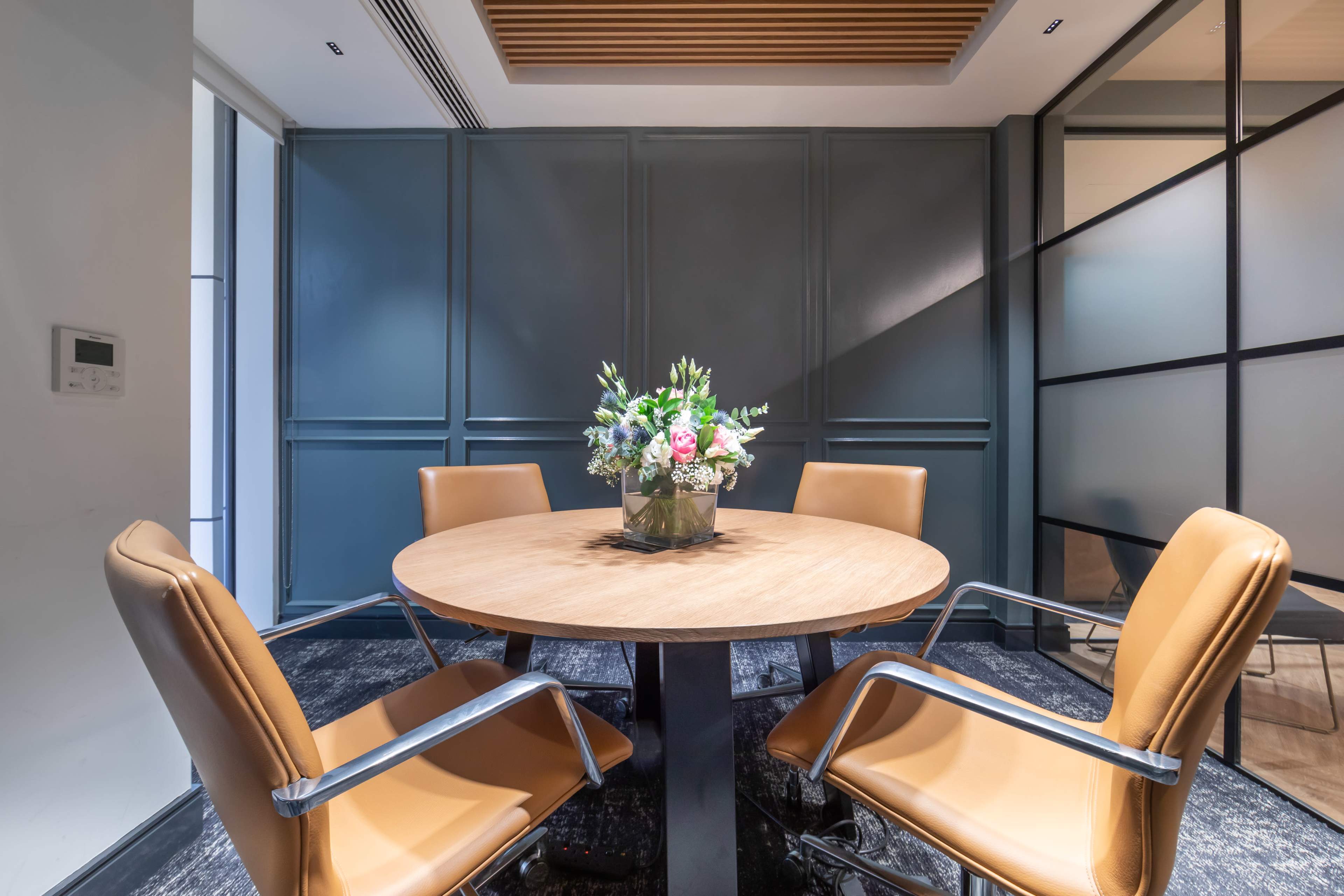 A round wooden table is centered in a meeting room with four brown leather chairs and a floral arrangement in the middle.