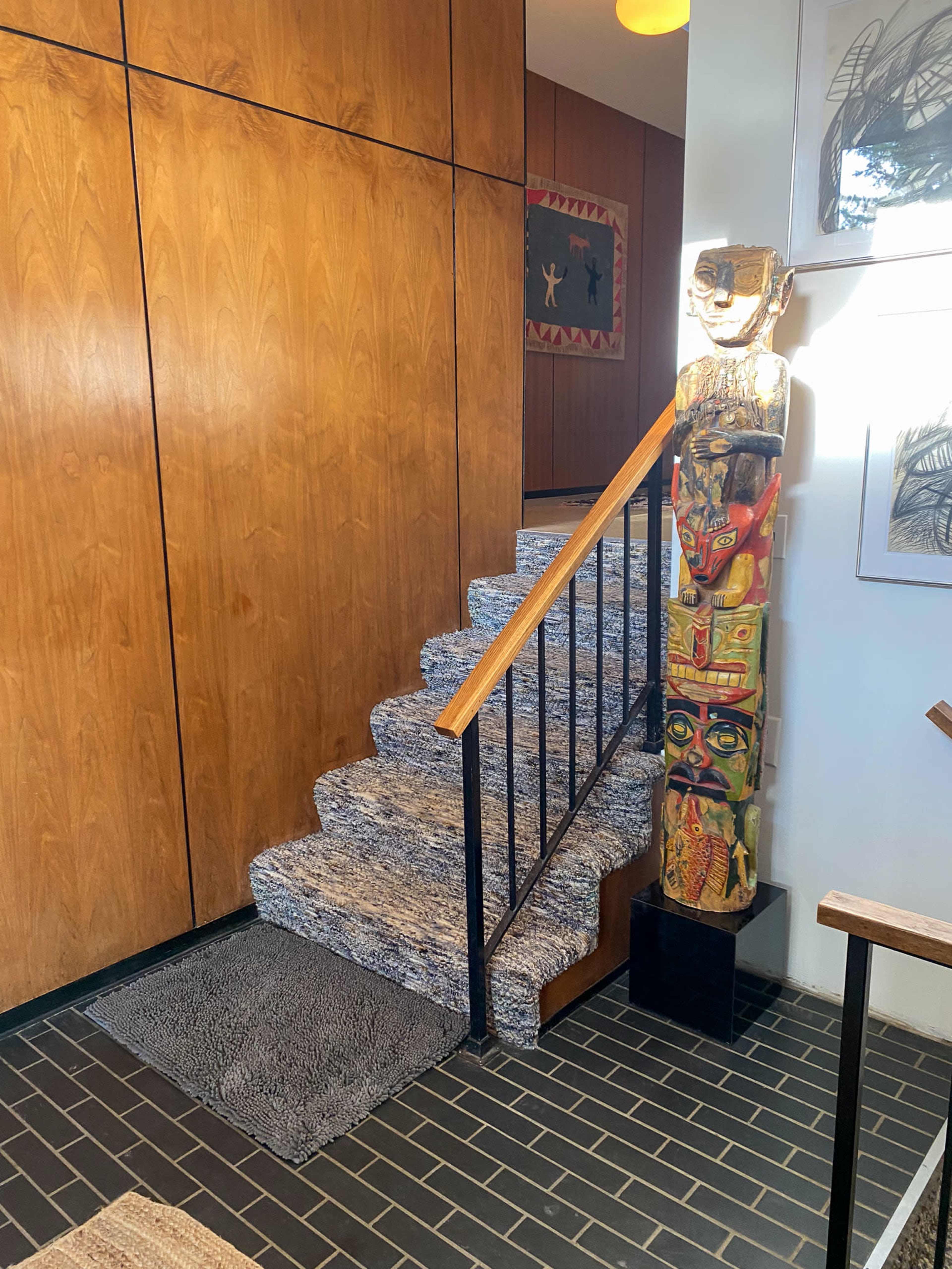 The image shows a staircase with a textured carpet leading up, adjacent to a wooden wall, with a colorful totem sculpture positioned beside the stairs.