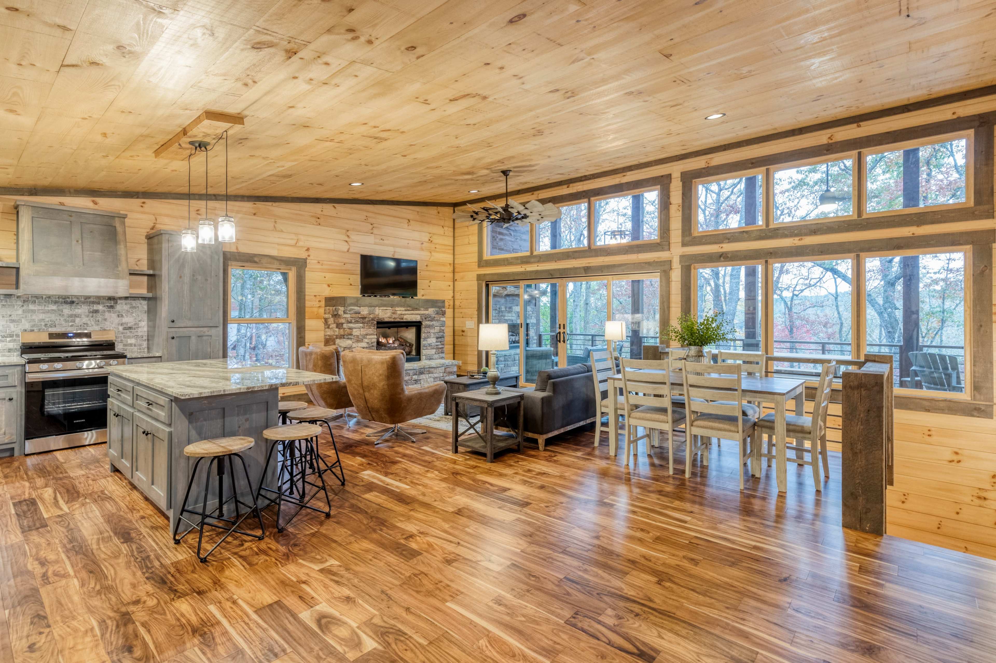 A modern cabin interior with wooden walls and floors, featuring a kitchen, dining area, and living space with large windows overlooking trees.