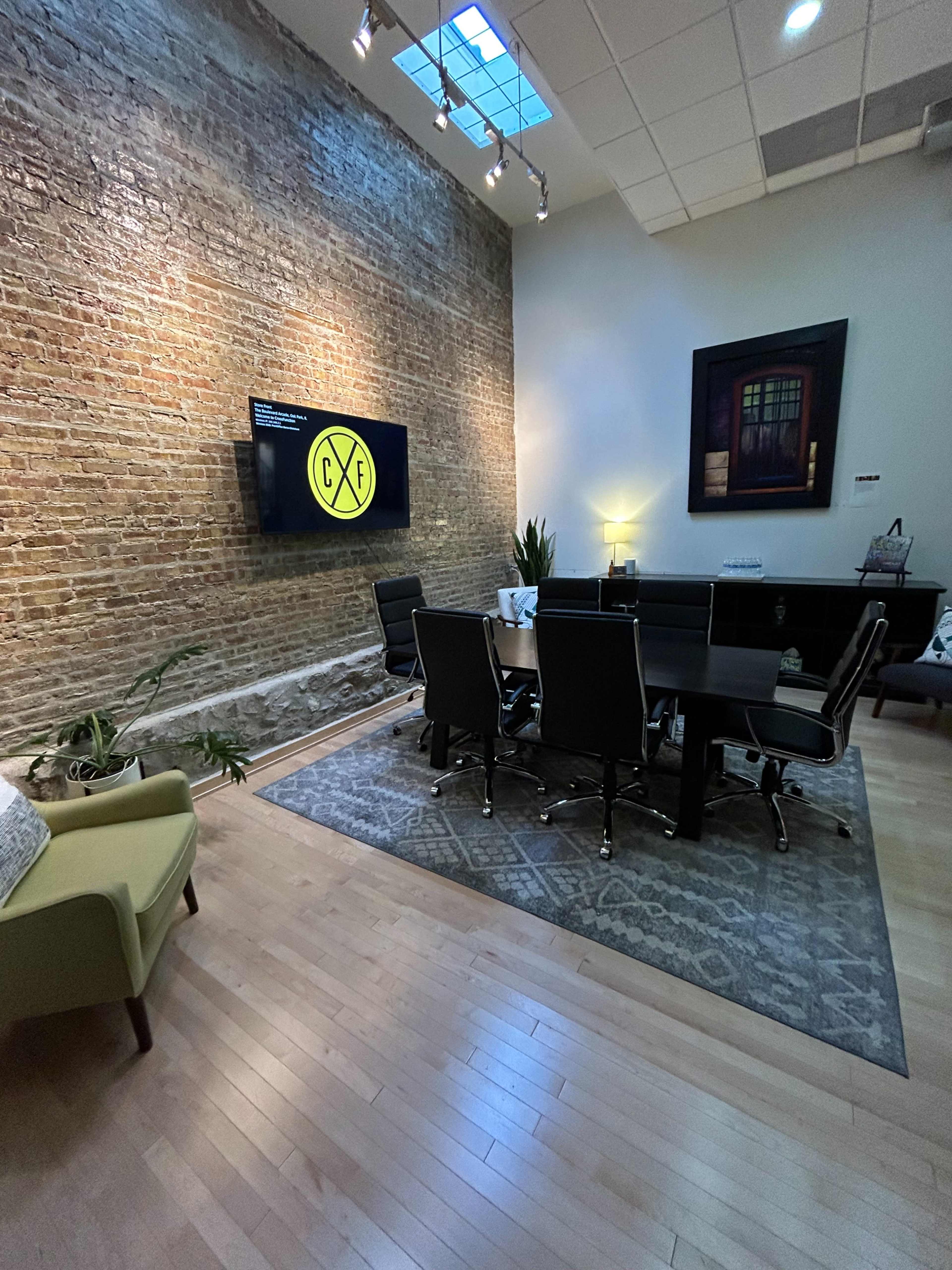 A modern conference room with a long table, black chairs, a brick wall, and a television mounted on the wall displaying a logo.