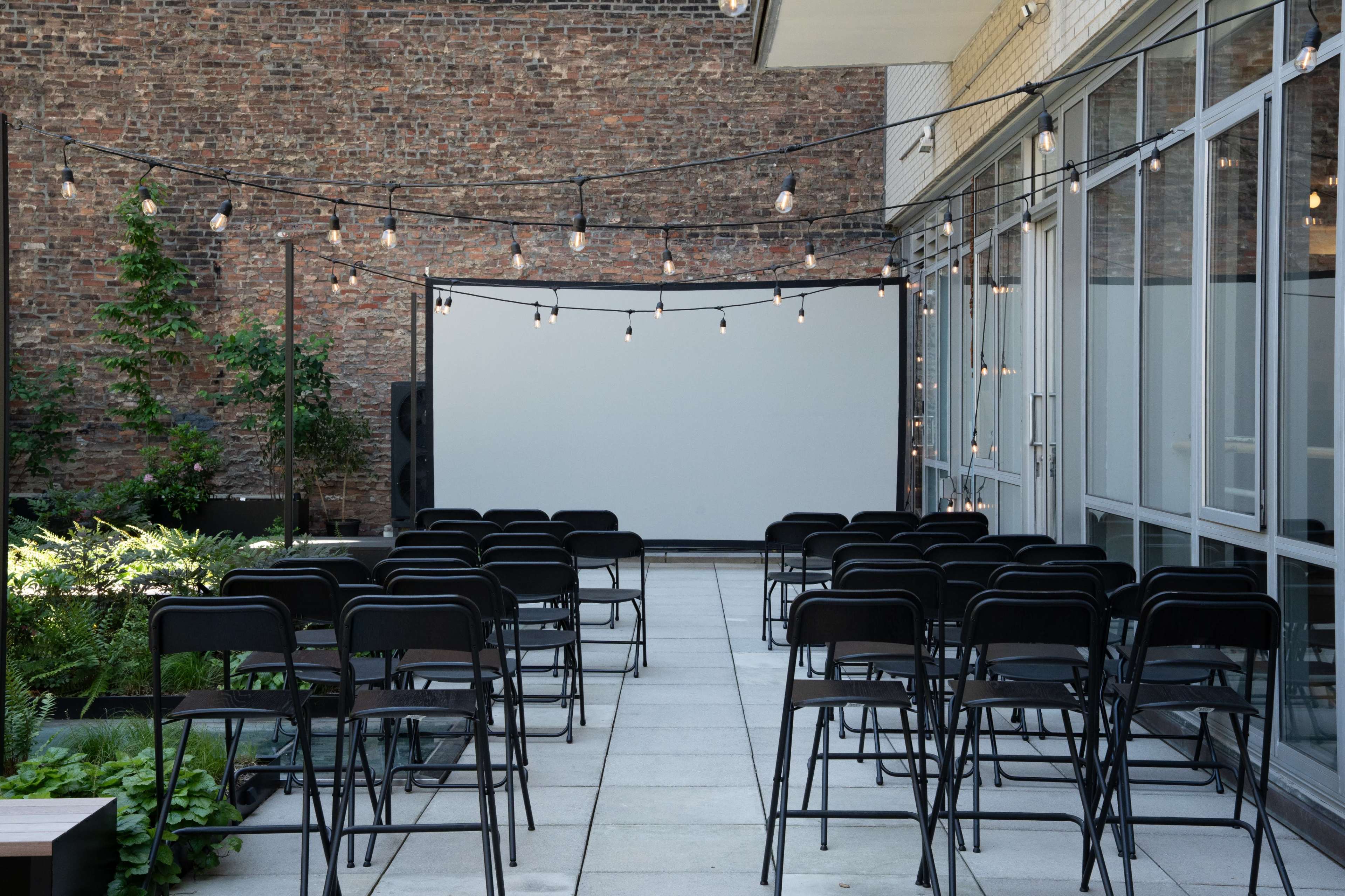 A courtyard is set up with rows of black chairs facing a blank screen, surrounded by greenery and strung lights overhead.