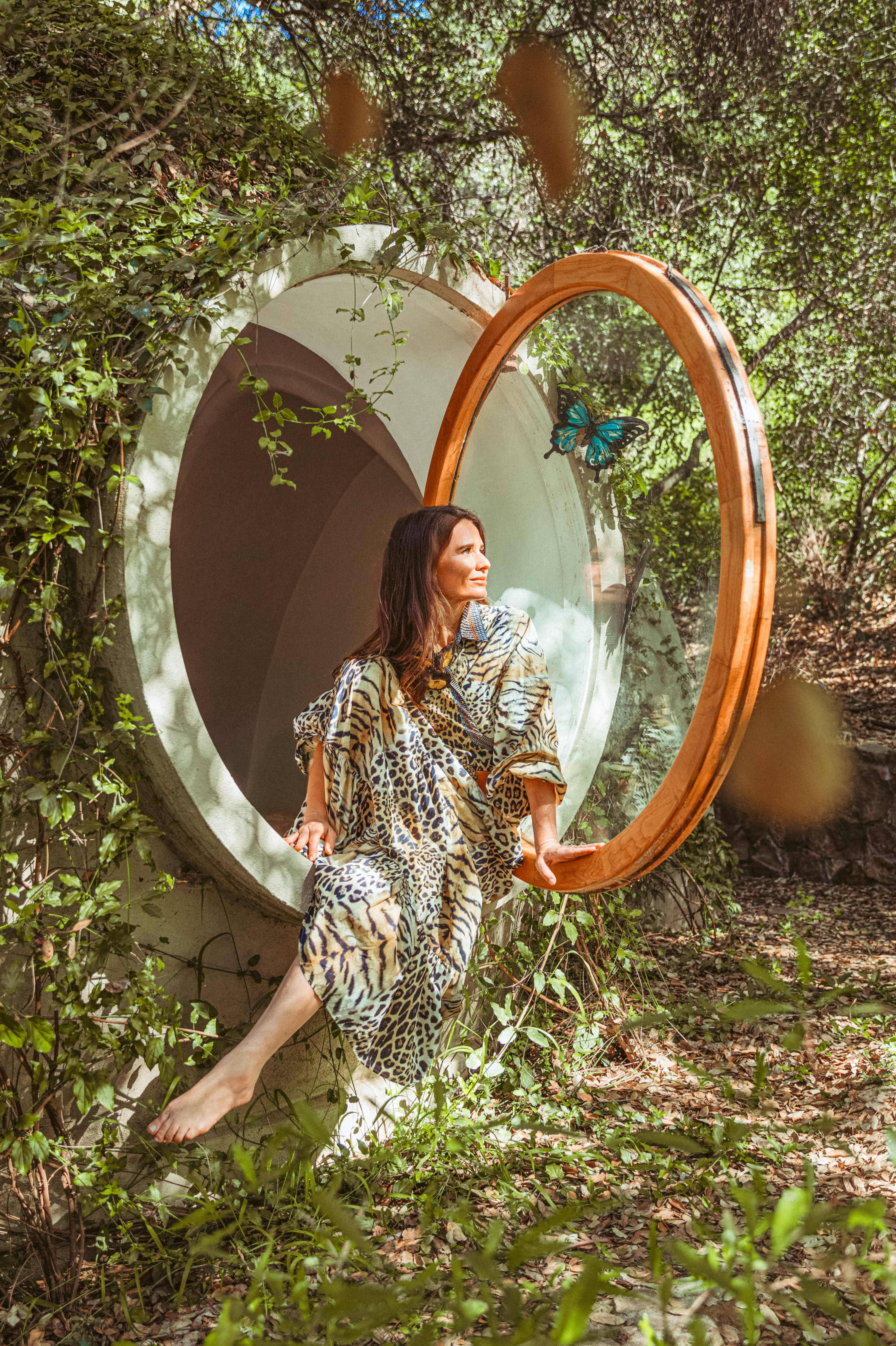 A woman sits gracefully on the edge of a circular window framed in wood, surrounded by lush greenery and natural light.