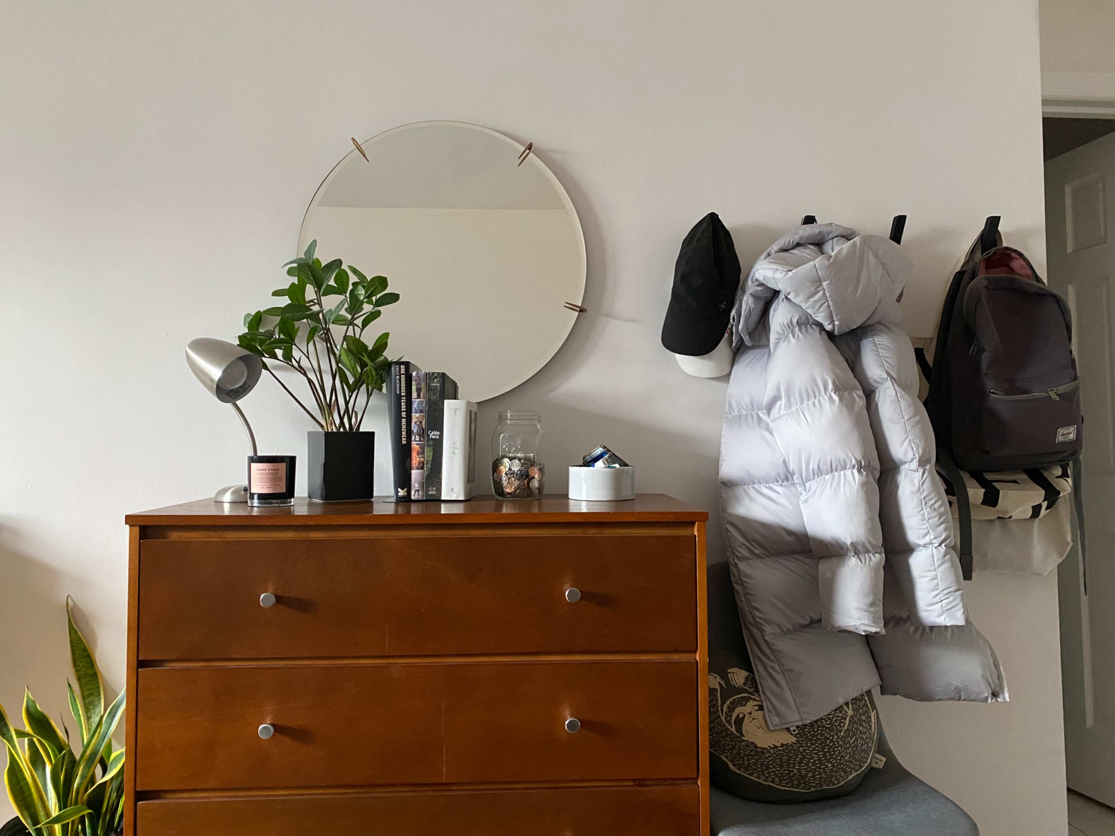 A wooden dresser with a mirror above it displays plants, books, and a lamp, while a grey puffer jacket, a hat, and two backpacks hang on the wall beside it.