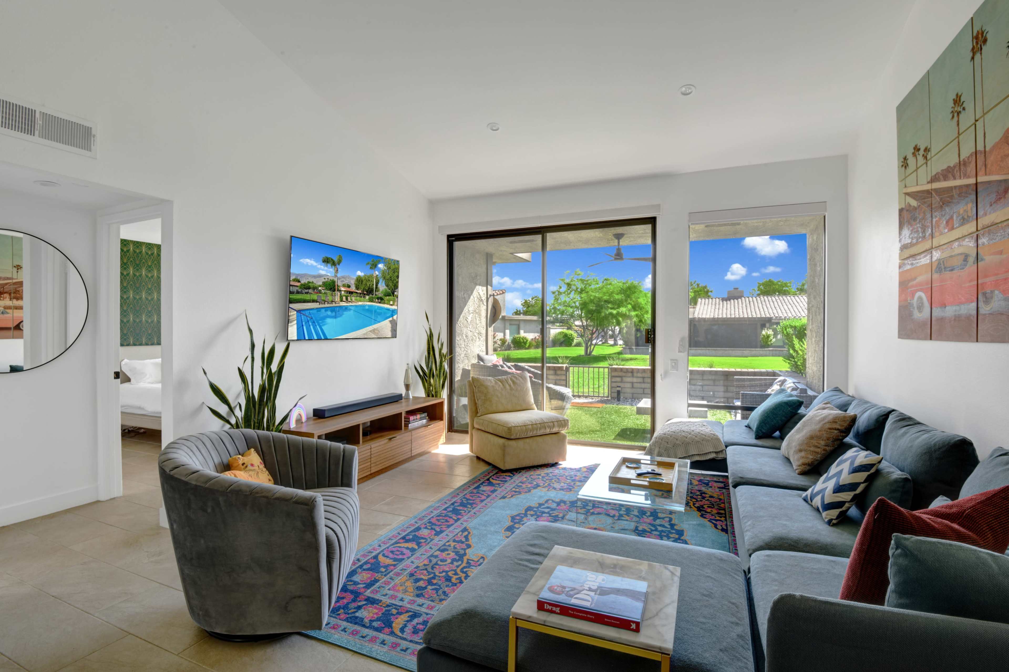 The living room features a sectional sofa, a coffee table, and a large window overlooking a green landscape with a pool visible outside.