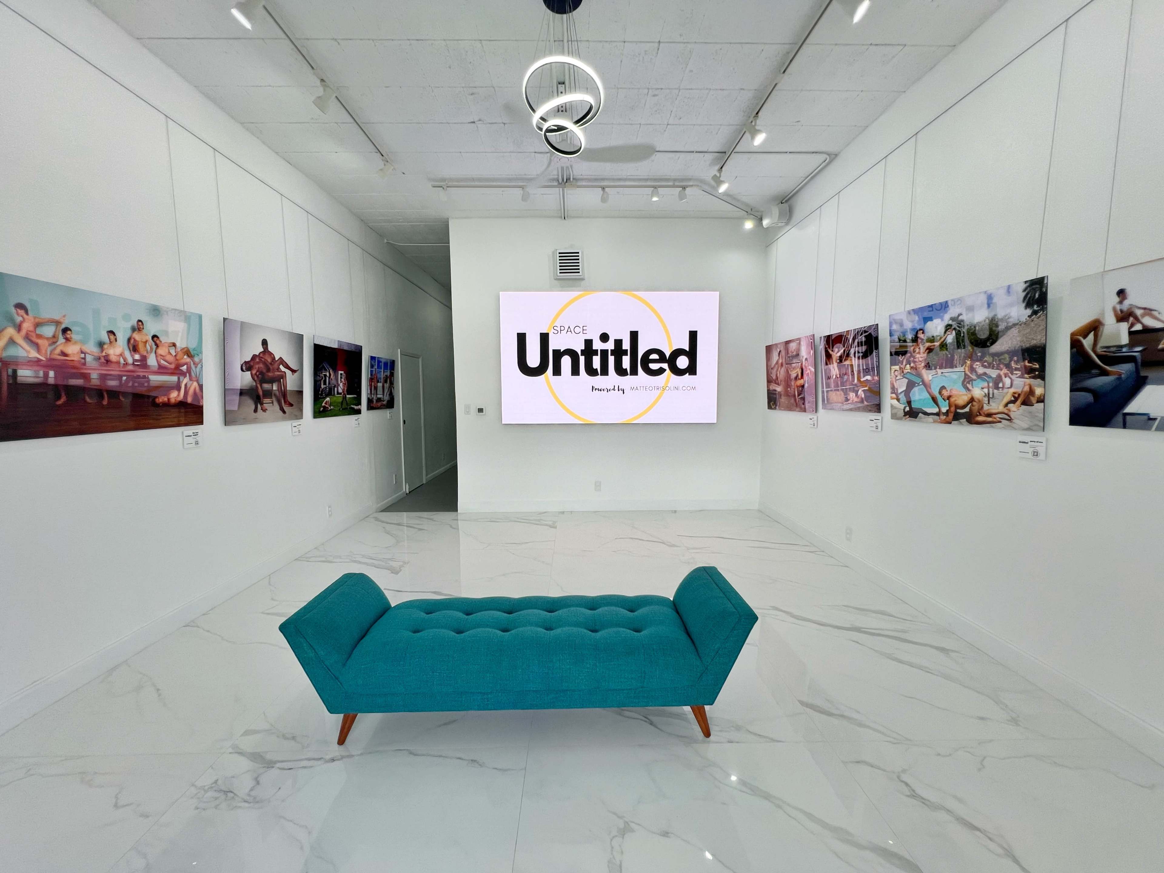The image shows a modern art gallery featuring a turquoise sofa in the center, surrounded by various artworks displayed on the walls, with a large screen displaying the word "Untitled."