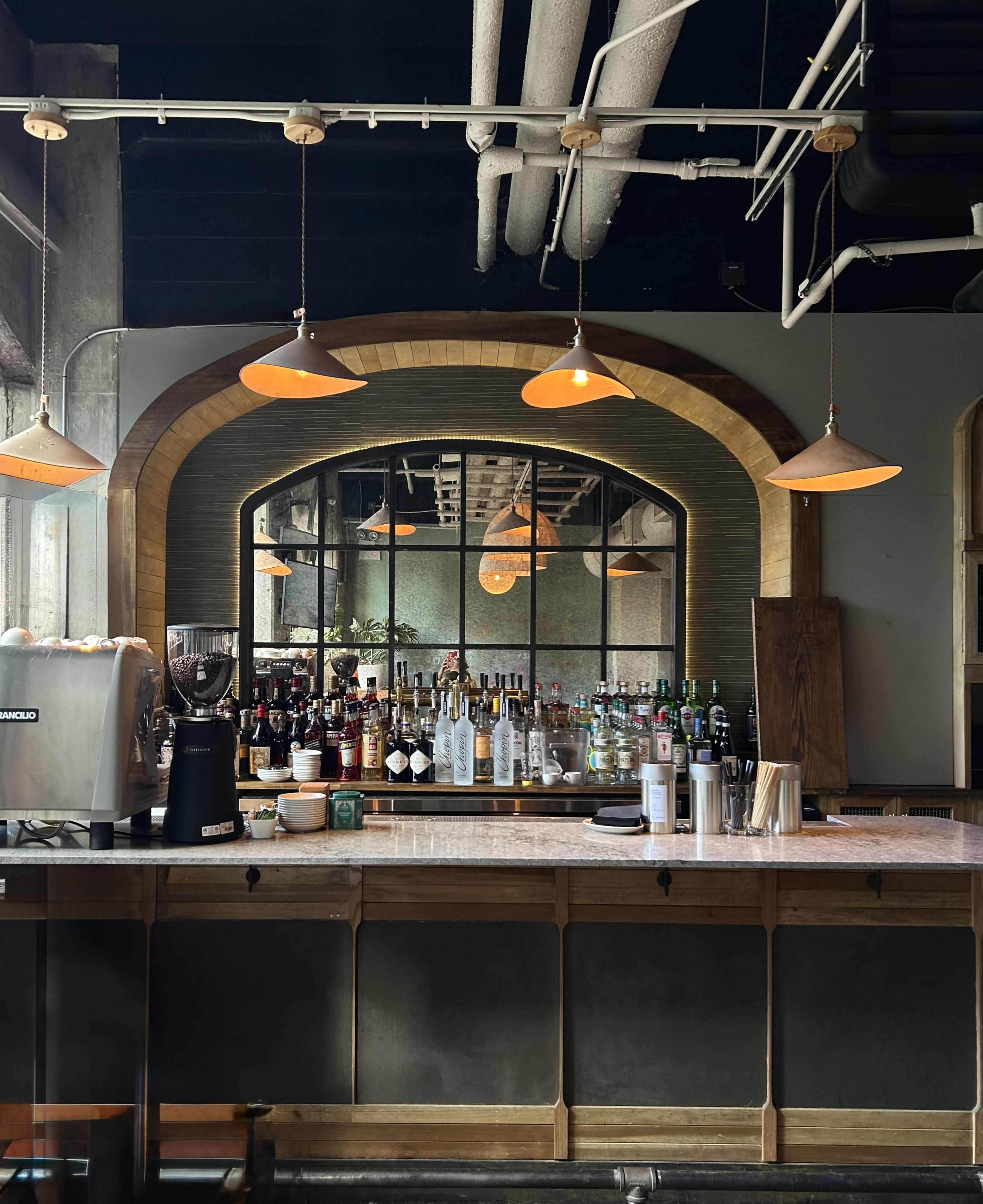 The image shows a bar area with a variety of bottles displayed on shelves behind the counter and warm pendant lights hanging above.
