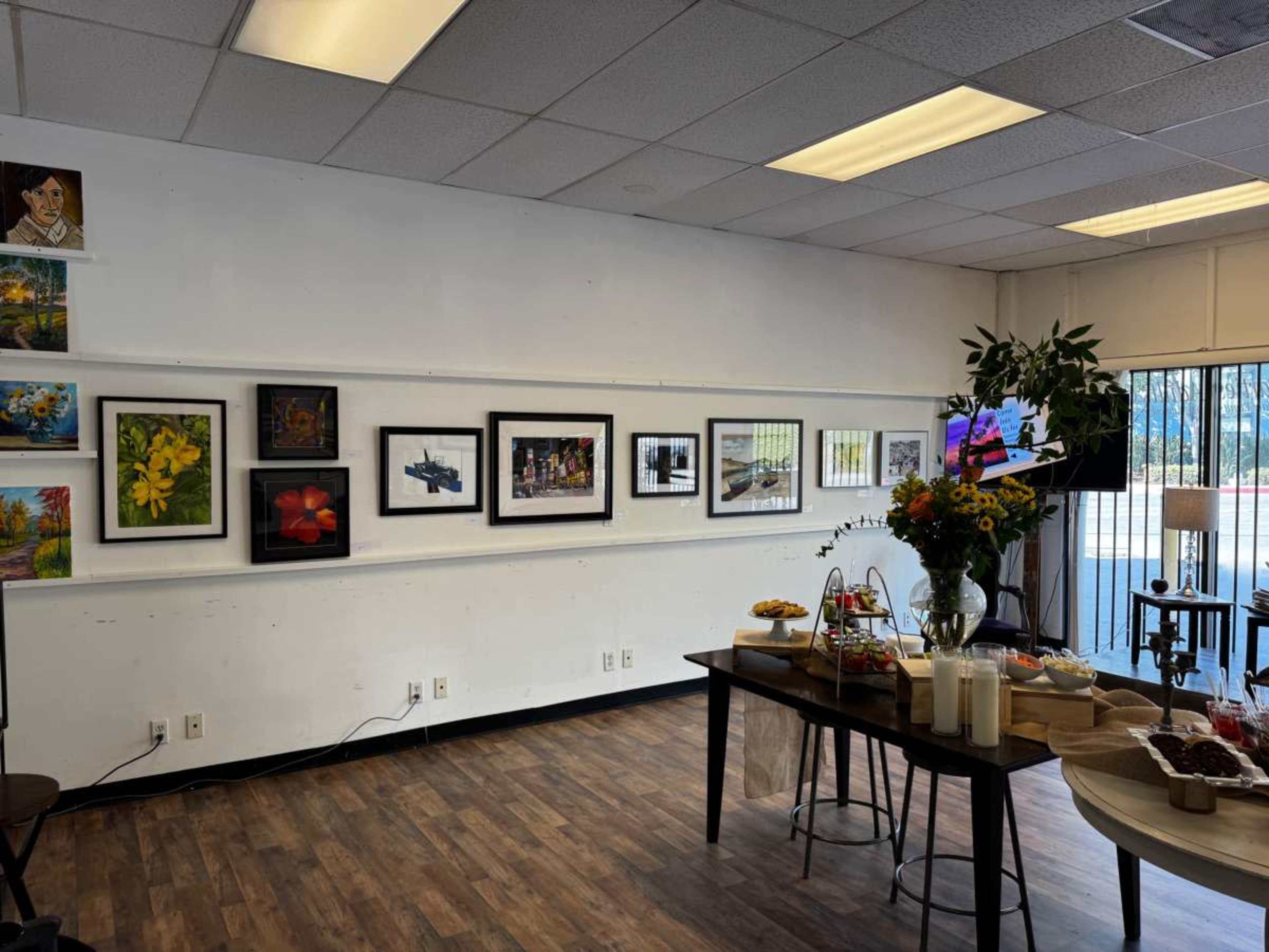 The image shows an art gallery interior featuring various framed artworks displayed on a white wall, with a table set up for refreshments in the foreground.