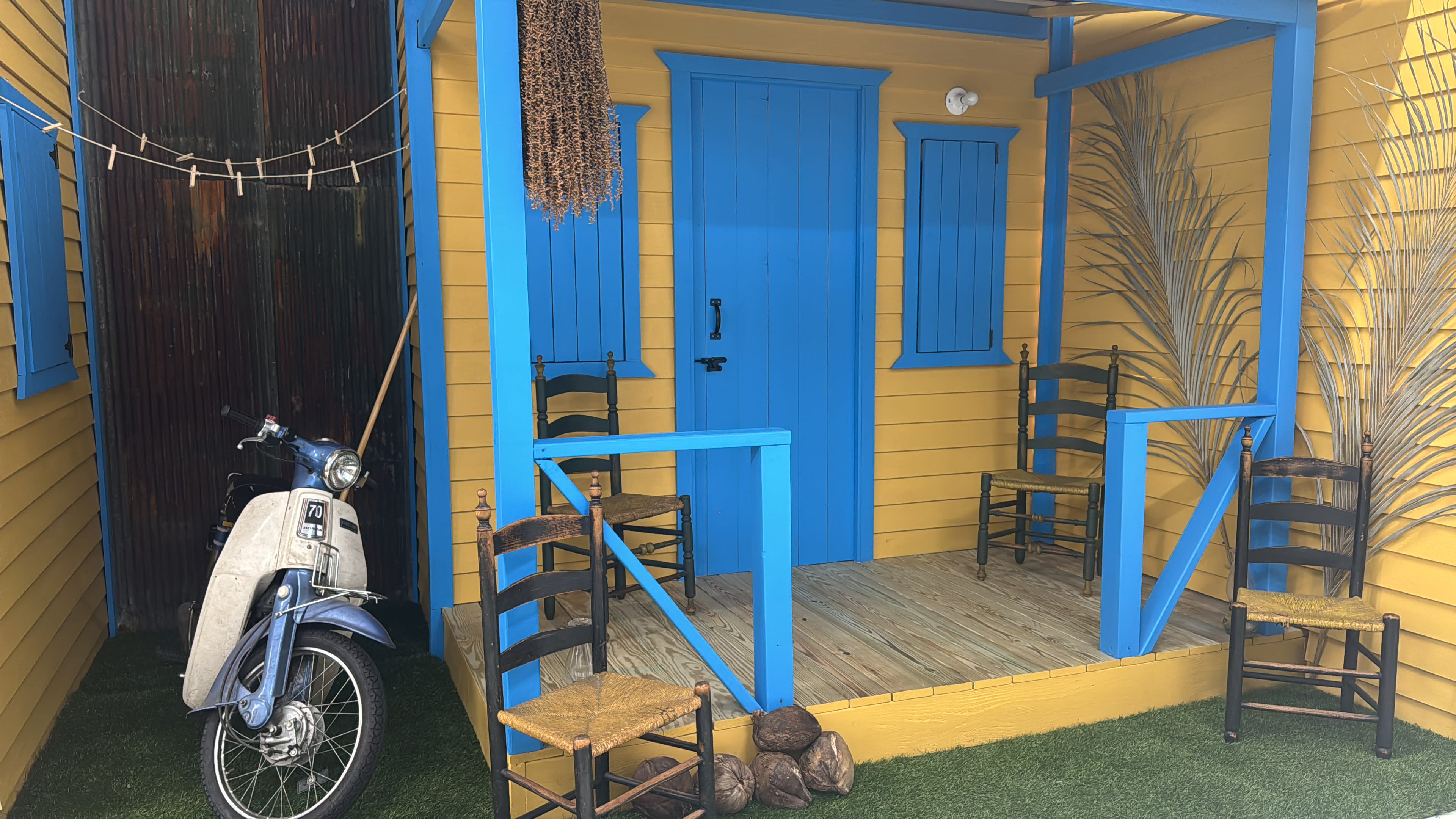The image shows a vibrant yellow house with a blue porch, featuring a vintage scooter and several wooden chairs arranged around a wooden deck.