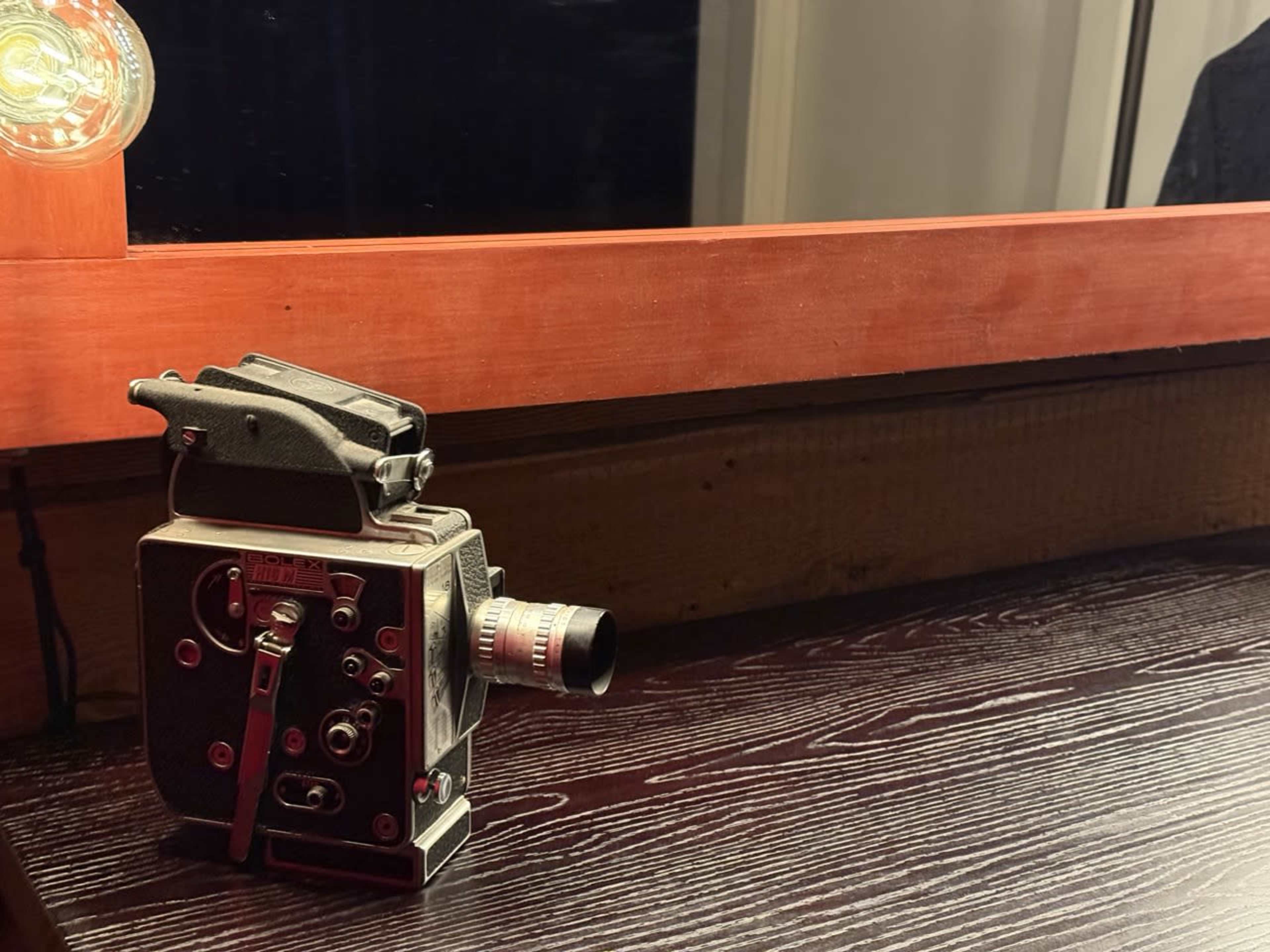 A vintage film camera is placed on a dark wooden surface next to a mirror reflecting warm light.