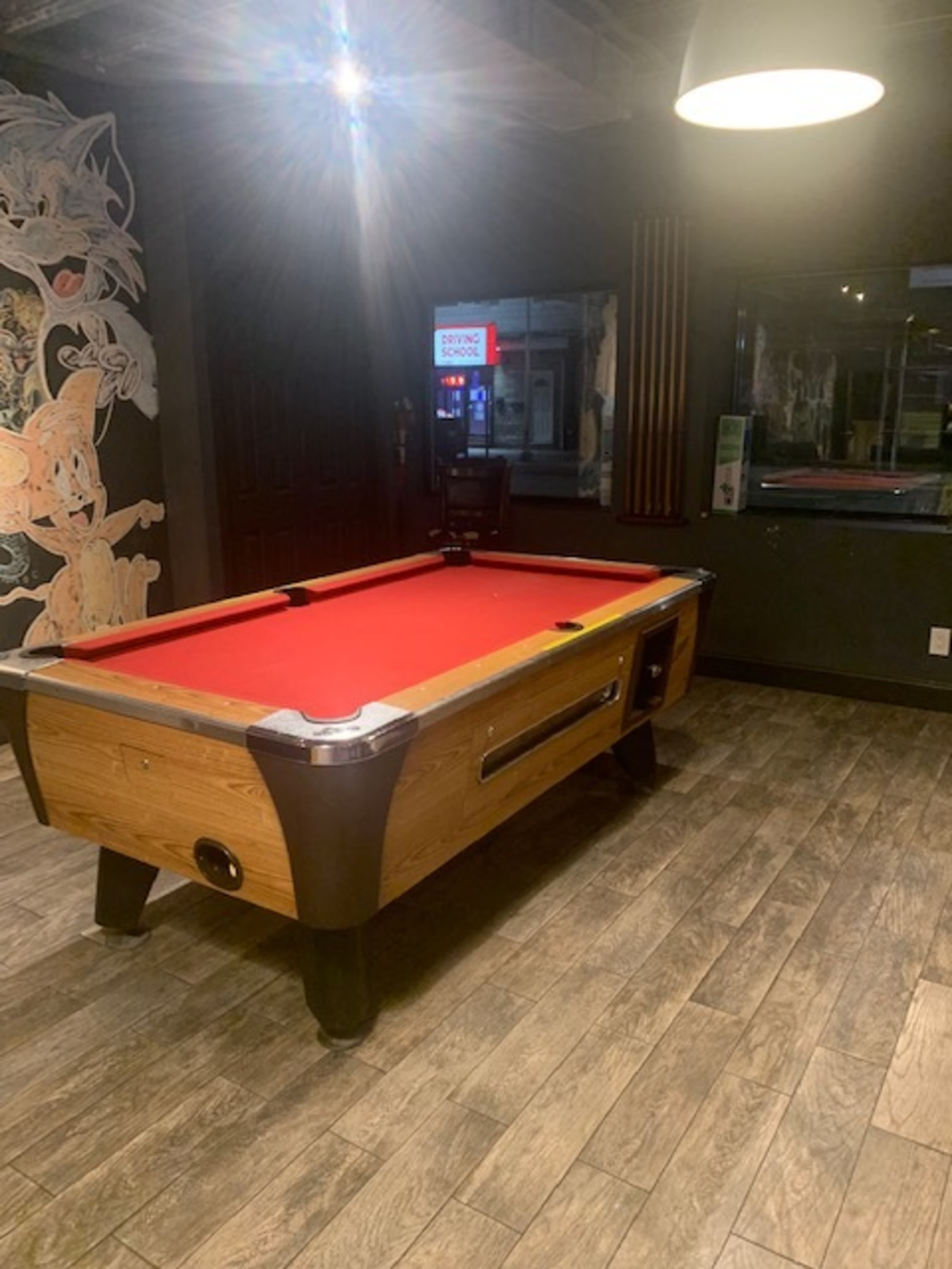 A pool table with a red felt surface located in a dimly lit room with wooden flooring and a mural on the wall.