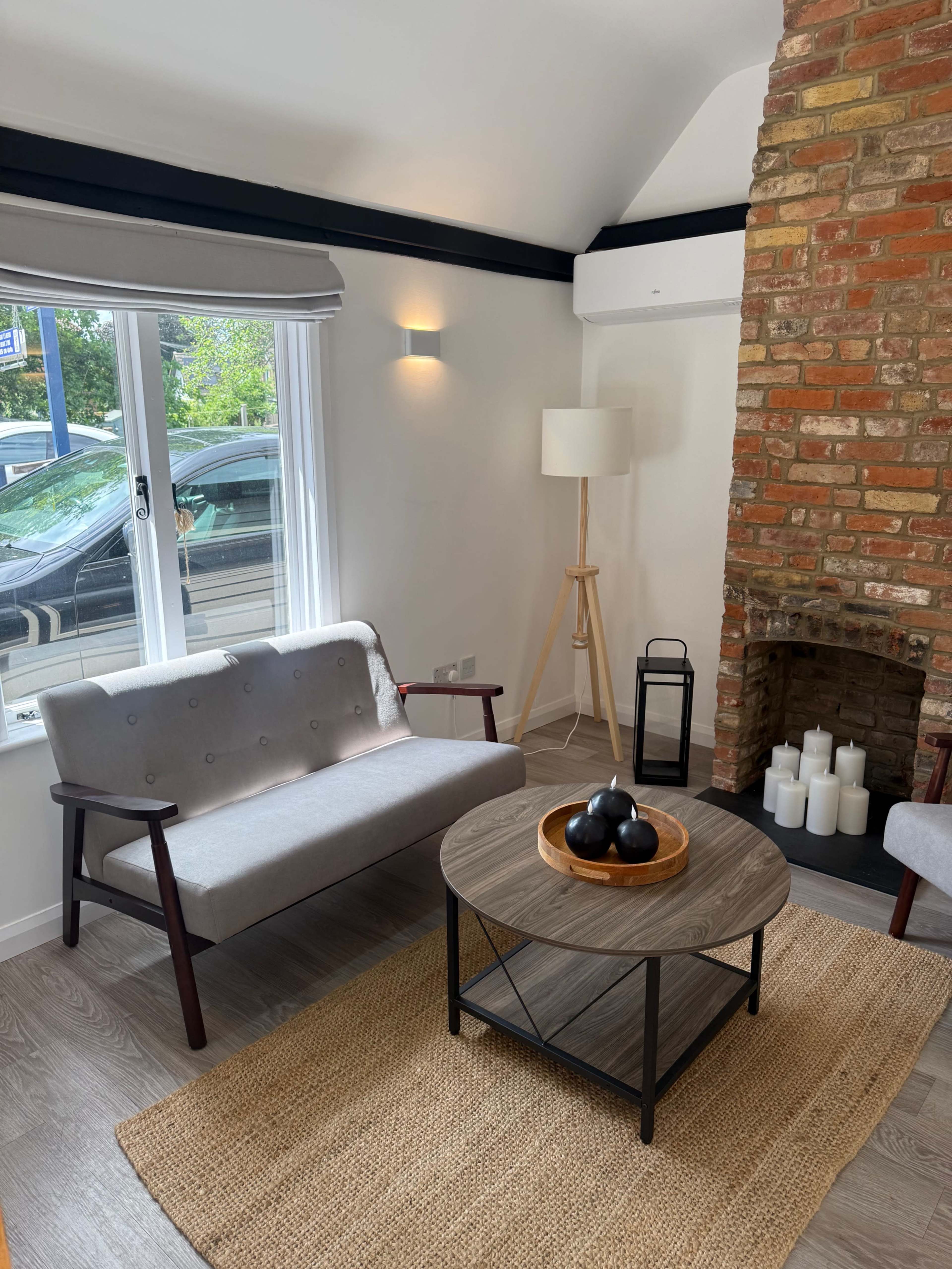 The image depicts a modern living room featuring a gray sofa, a round coffee table with decorative items, a floor lamp, and a brick fireplace surrounded by white candles.