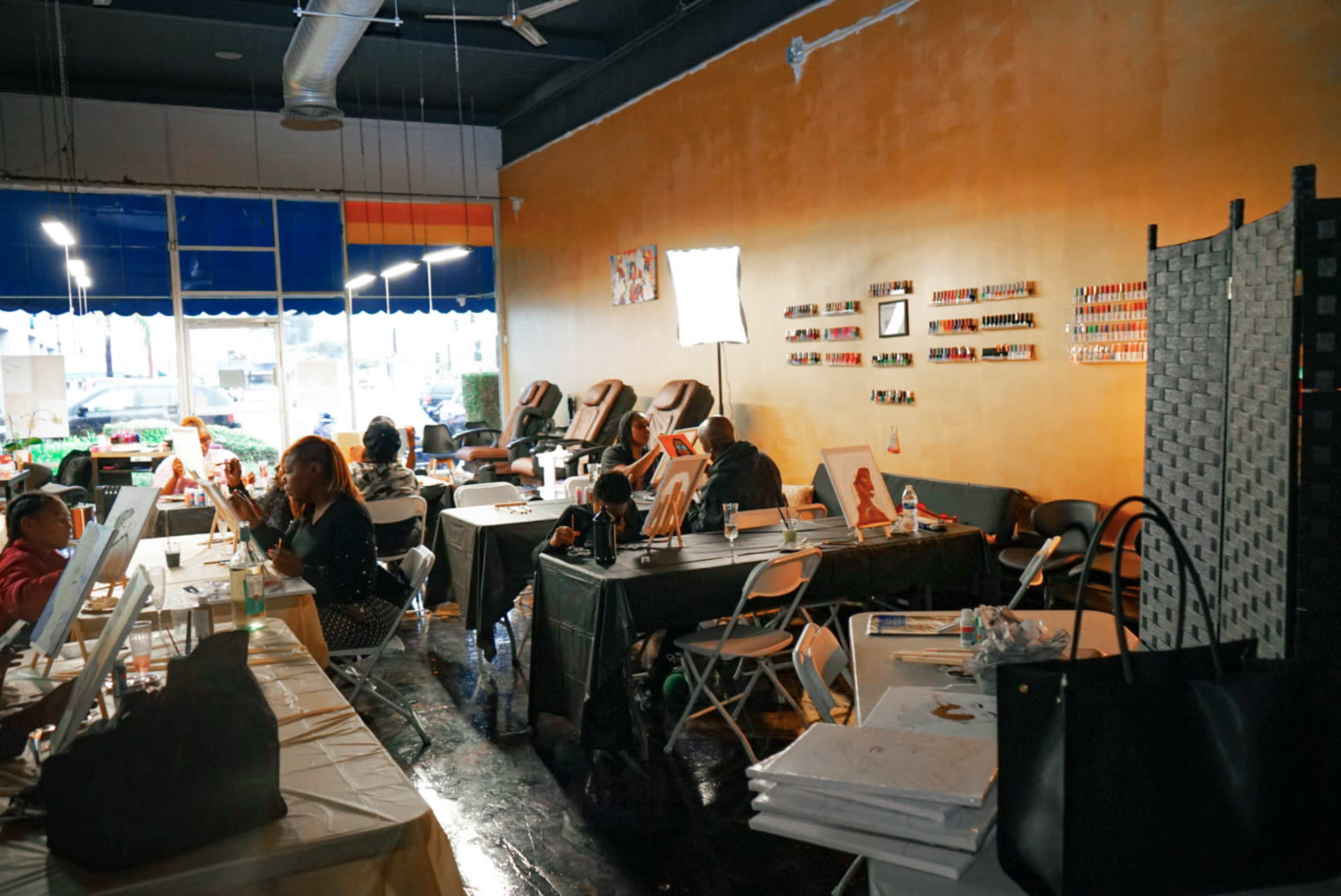 A brightly lit nail salon with customers seated at tables, manicure stations, and a display of nail polish on the walls.