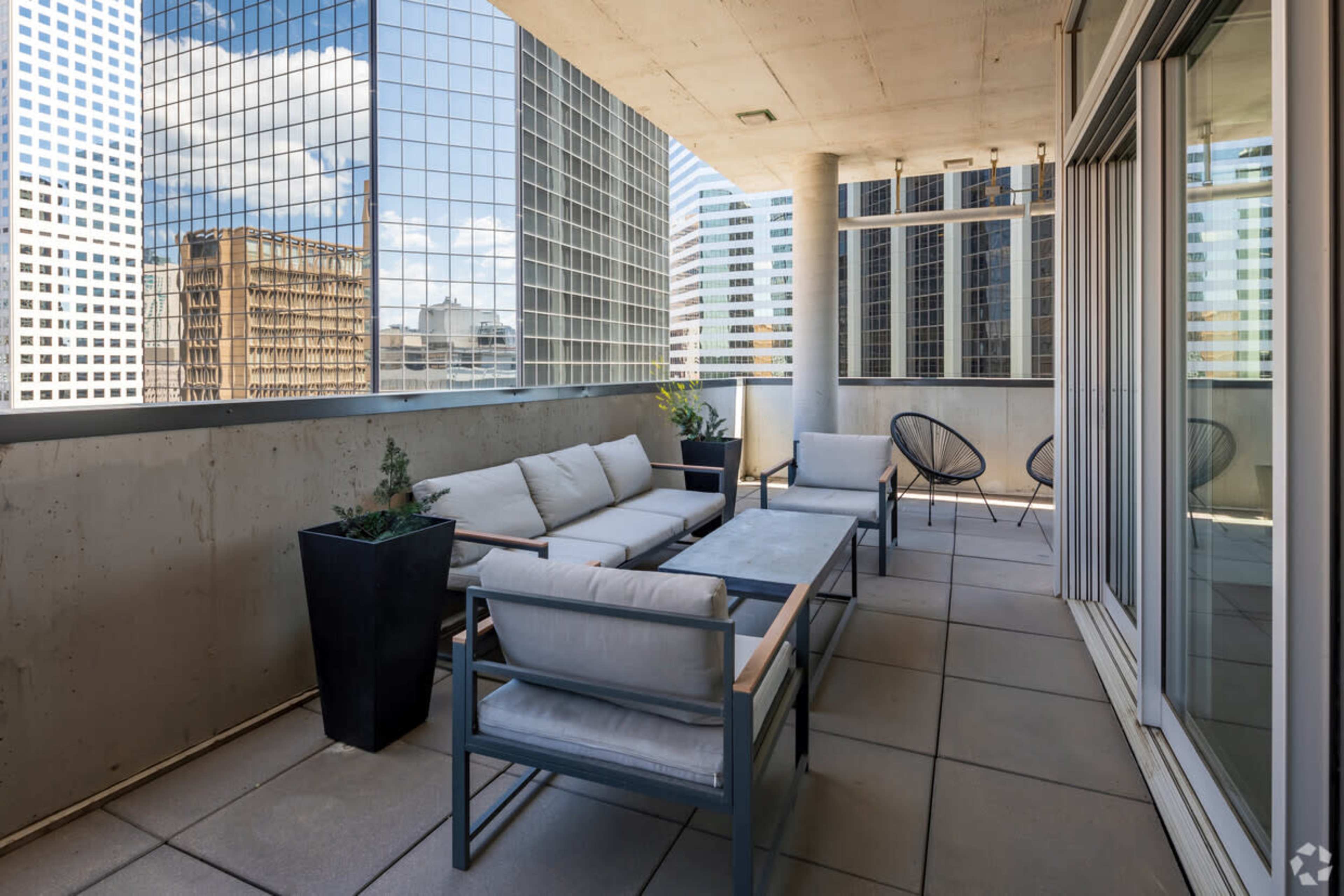 A modern balcony features a gray seating area with cushions, a rectangular table, and black planters, overlooking tall glass buildings.