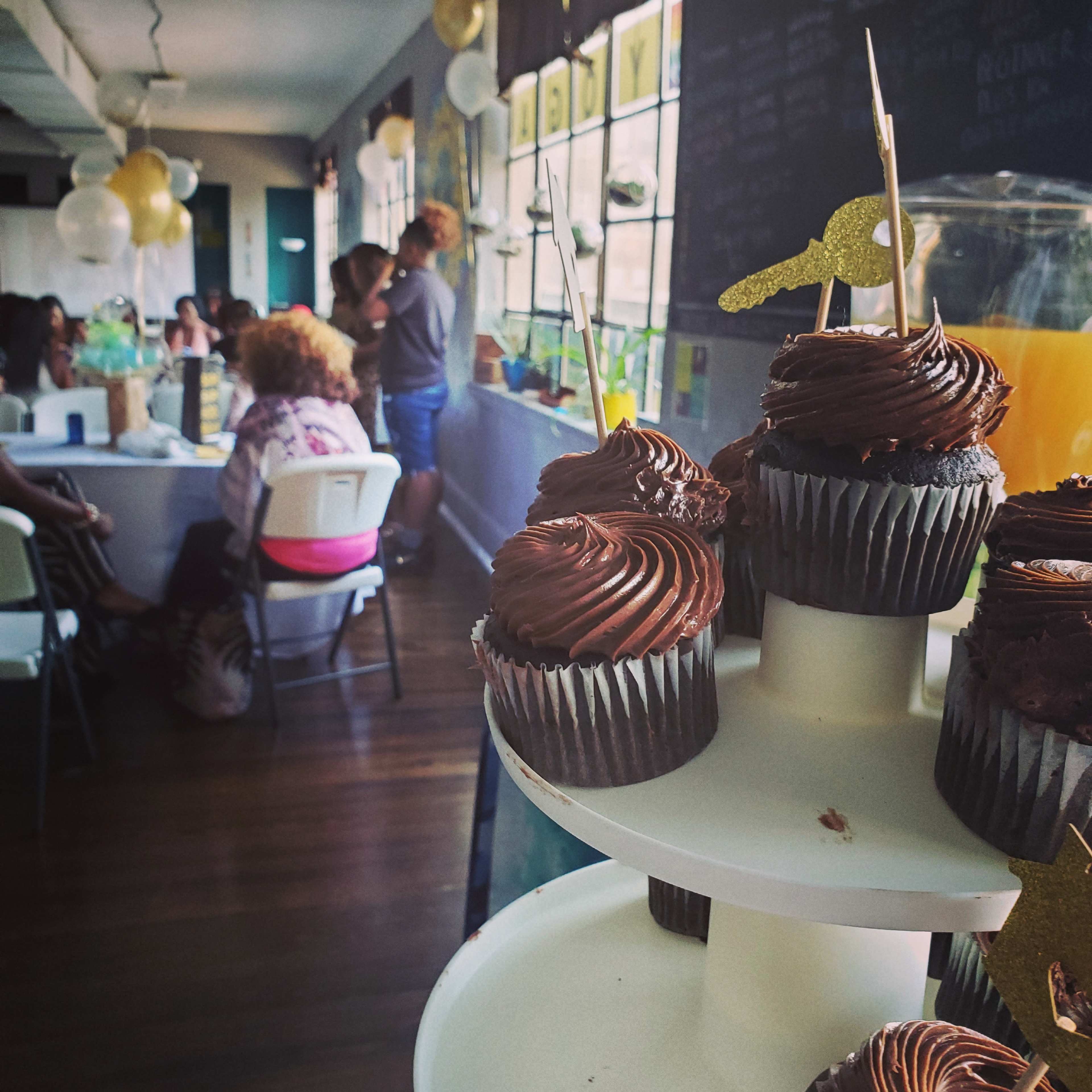 A tiered stand of chocolate cupcakes with decorative toothpicks is positioned in the foreground, while a gathering of people is visible in the background.