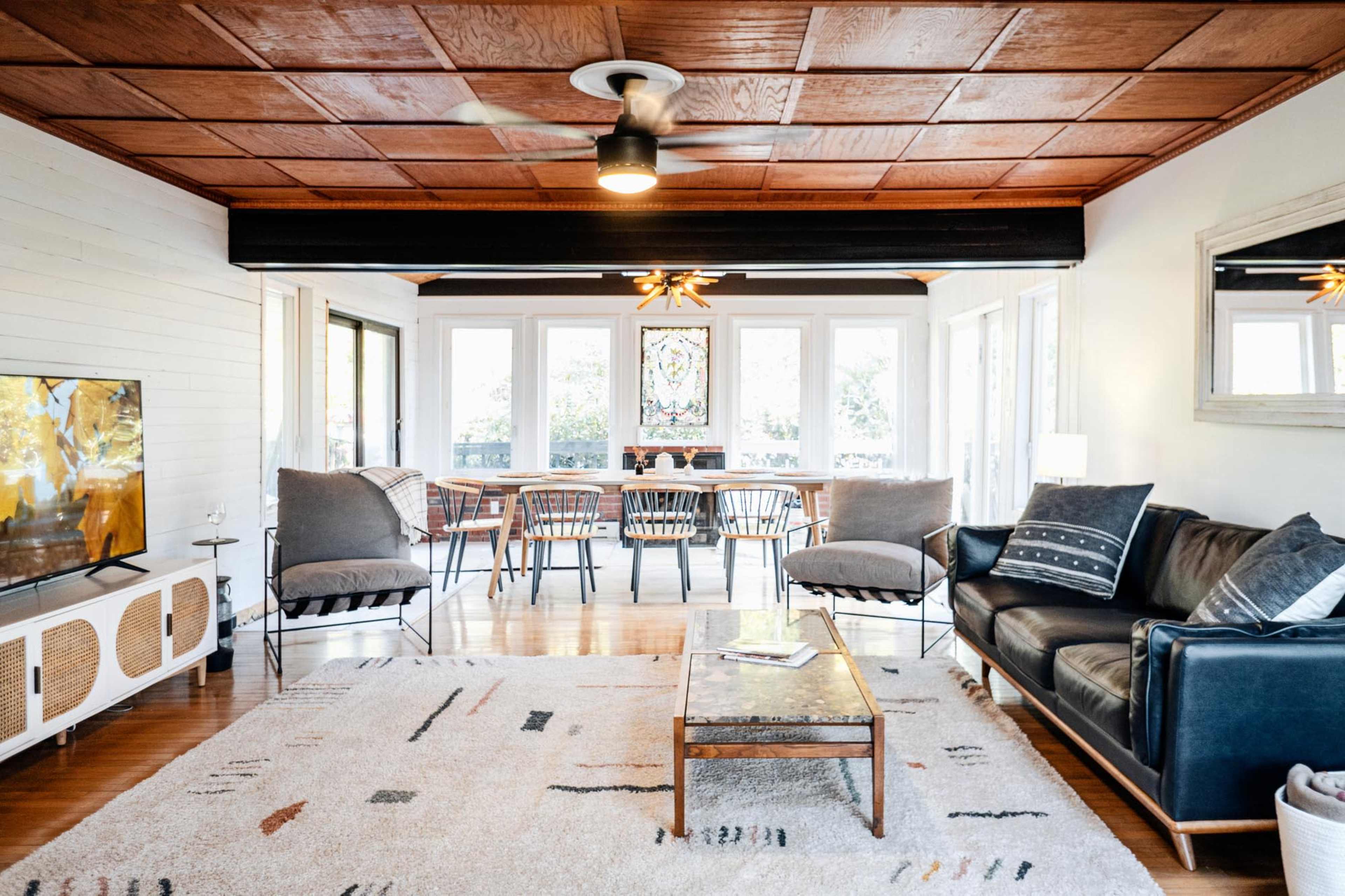 A well-furnished living room features a sofa, coffee table, and a television, with a dining area visible in the background through large windows.